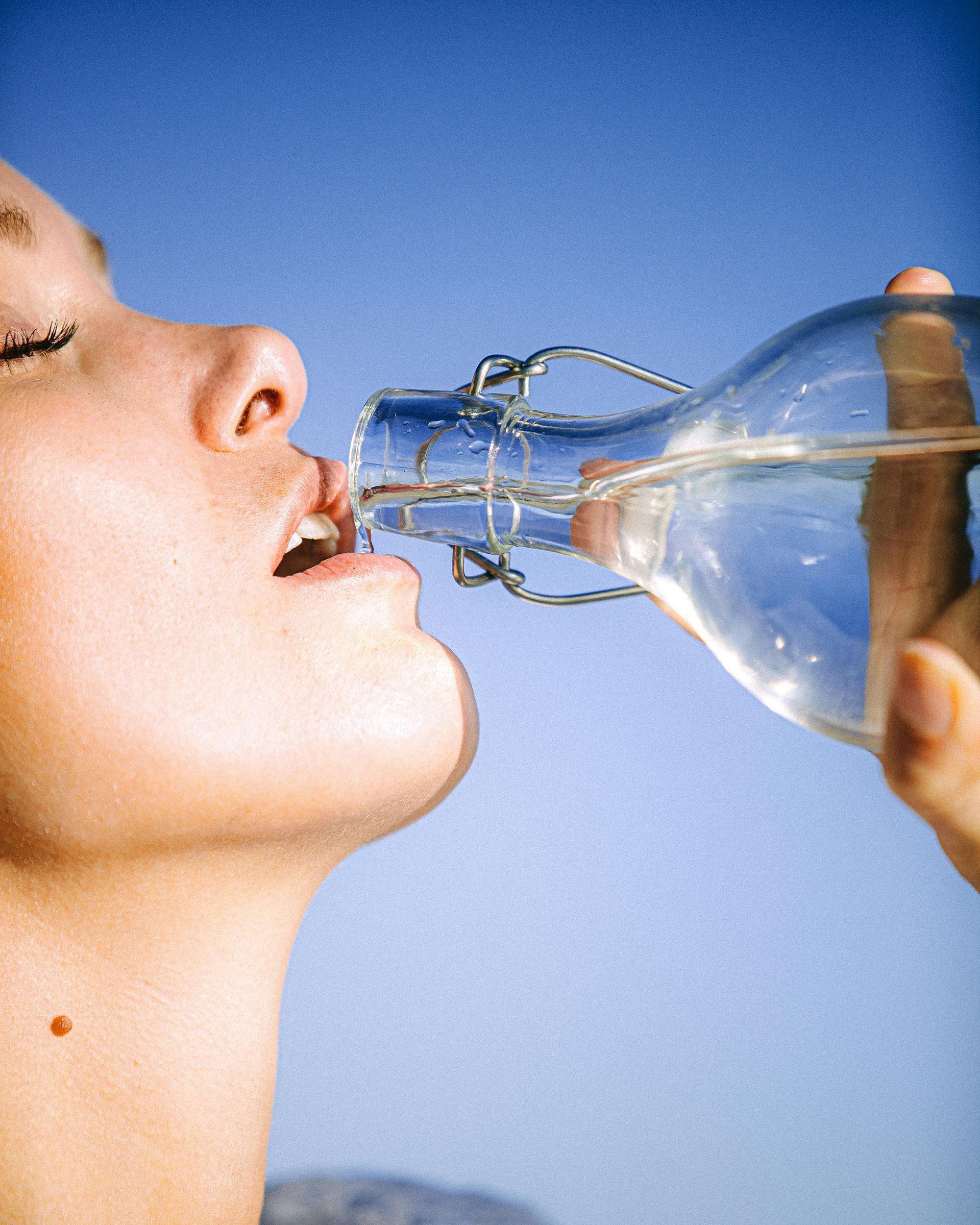 Woman drinking water from a clear glass bottle against a blue sky.