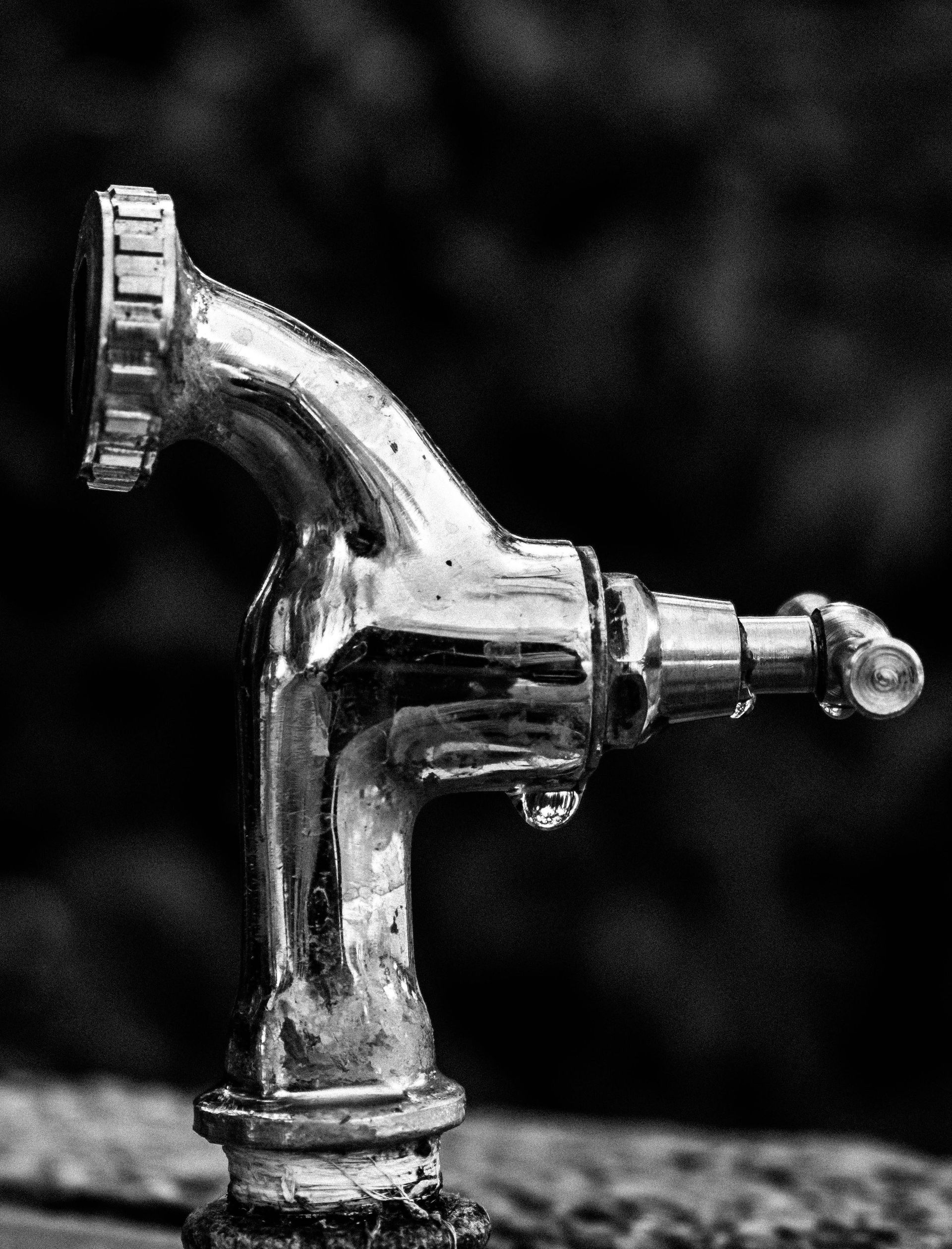A chrome water tap with a single water droplet hanging at the spout, dark background.