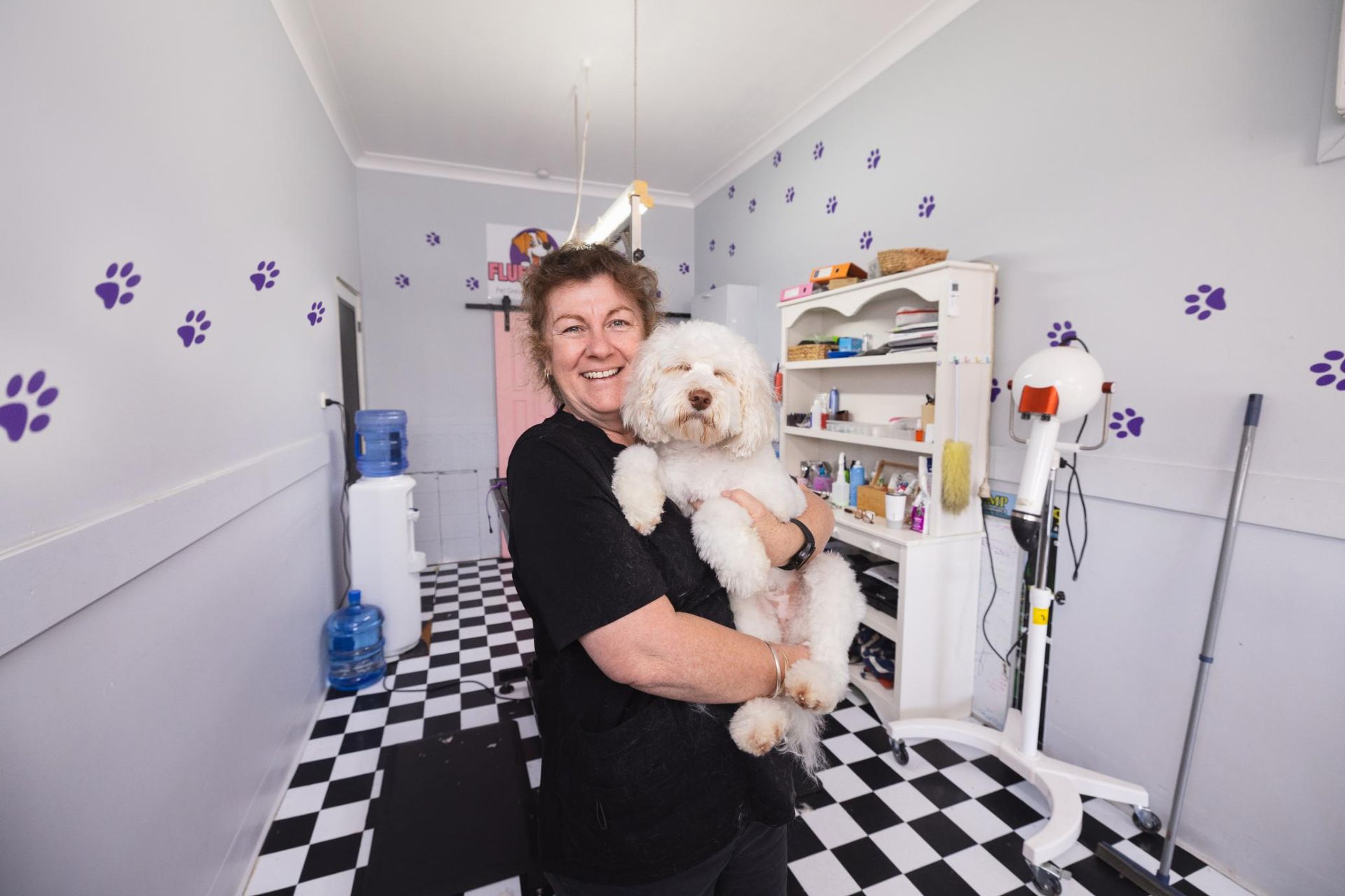 A Woman is Holding a Small White Dog in Her Arms in a Room — Fluffy Paws Pet Grooming In Wingham, NSW