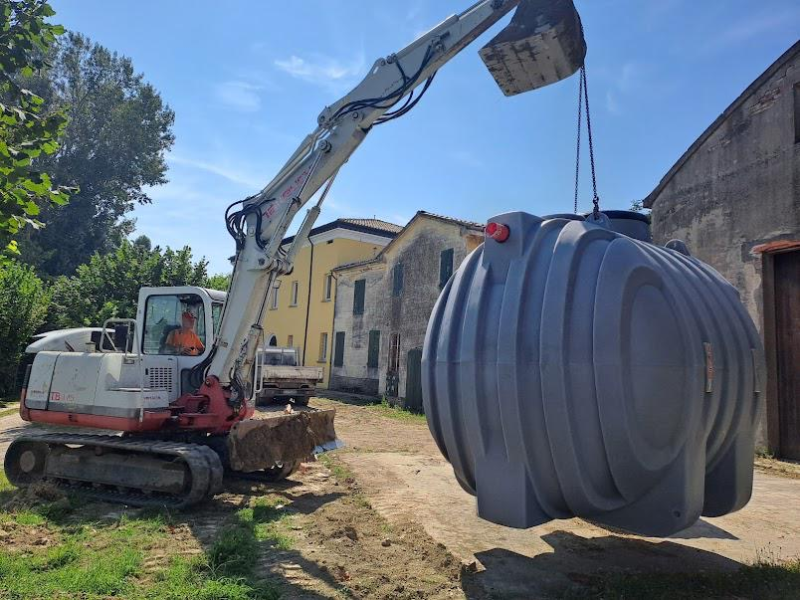 Un escavatore solleva un grande serbatoio sotterraneo in plastica grigia e nervata in una zona rurale vicino ad alcuni edifici.