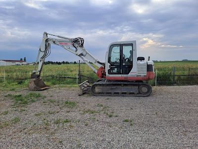 Un miniescavatore bianco e rosso parcheggiato su un piazzale ghiaioso di fronte a un campo, sotto un cielo nuvoloso.