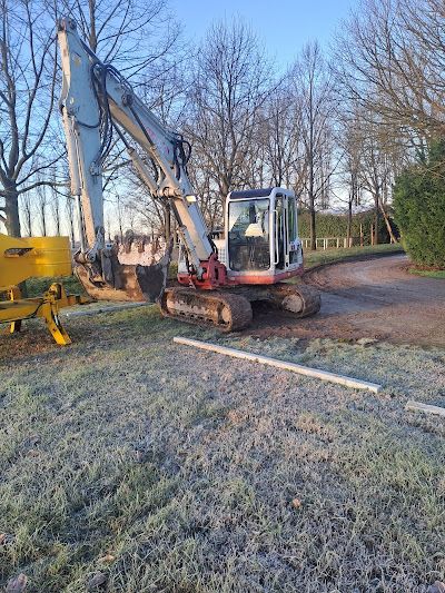 In una giornata di sole, un escavatore bianco è parcheggiato sul ciglio erboso accanto a un sentiero sterrato e a un macchinario giallo.