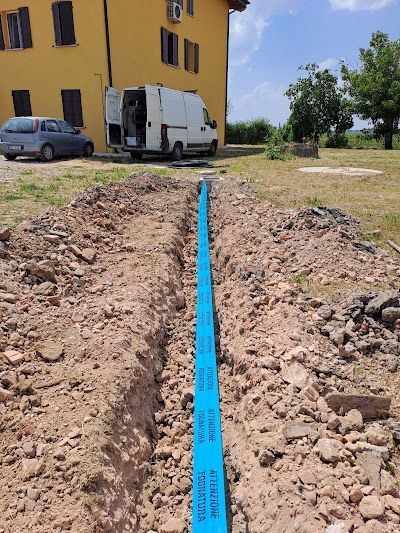 In un contesto rurale, un tubo di un blu brillante giace in una trincea di terra a cielo aperto, vicino a un edificio giallo e a un furgone da lavoro bianco.