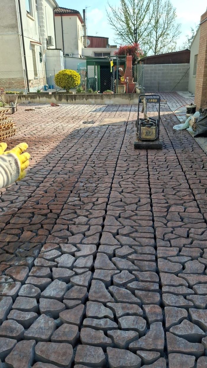 Un operaio utilizza una piastra vibrante su un patio appena posato, costituito da ciottoli irregolari di colore terroso, in un cortile.