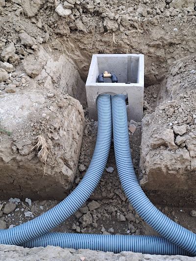 Due tubi flessibili blu entrano in una scatola di derivazione quadrata in cemento situata in una trincea di terra aperta.