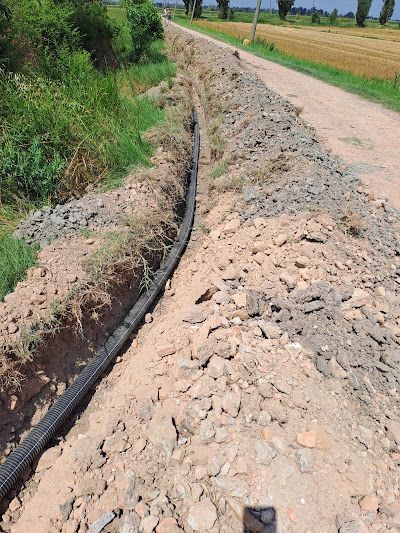 Un tubo nero per impianti elettrici giace in una trincea a cielo aperto lungo una strada sterrata di campagna, accanto a un campo.