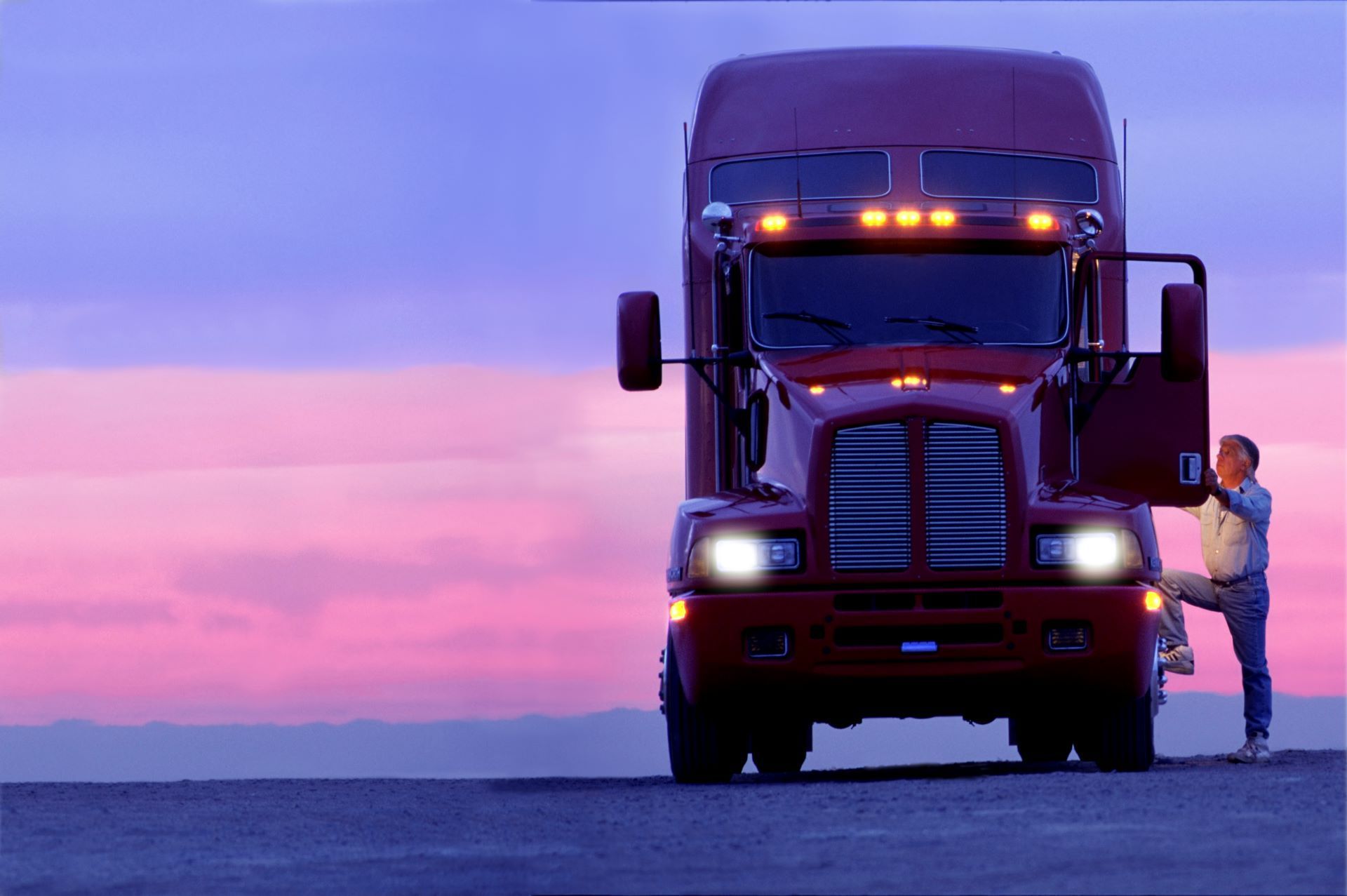 Red semi-truck at dusk with driver stepping out of cab. Purple and pink sky.