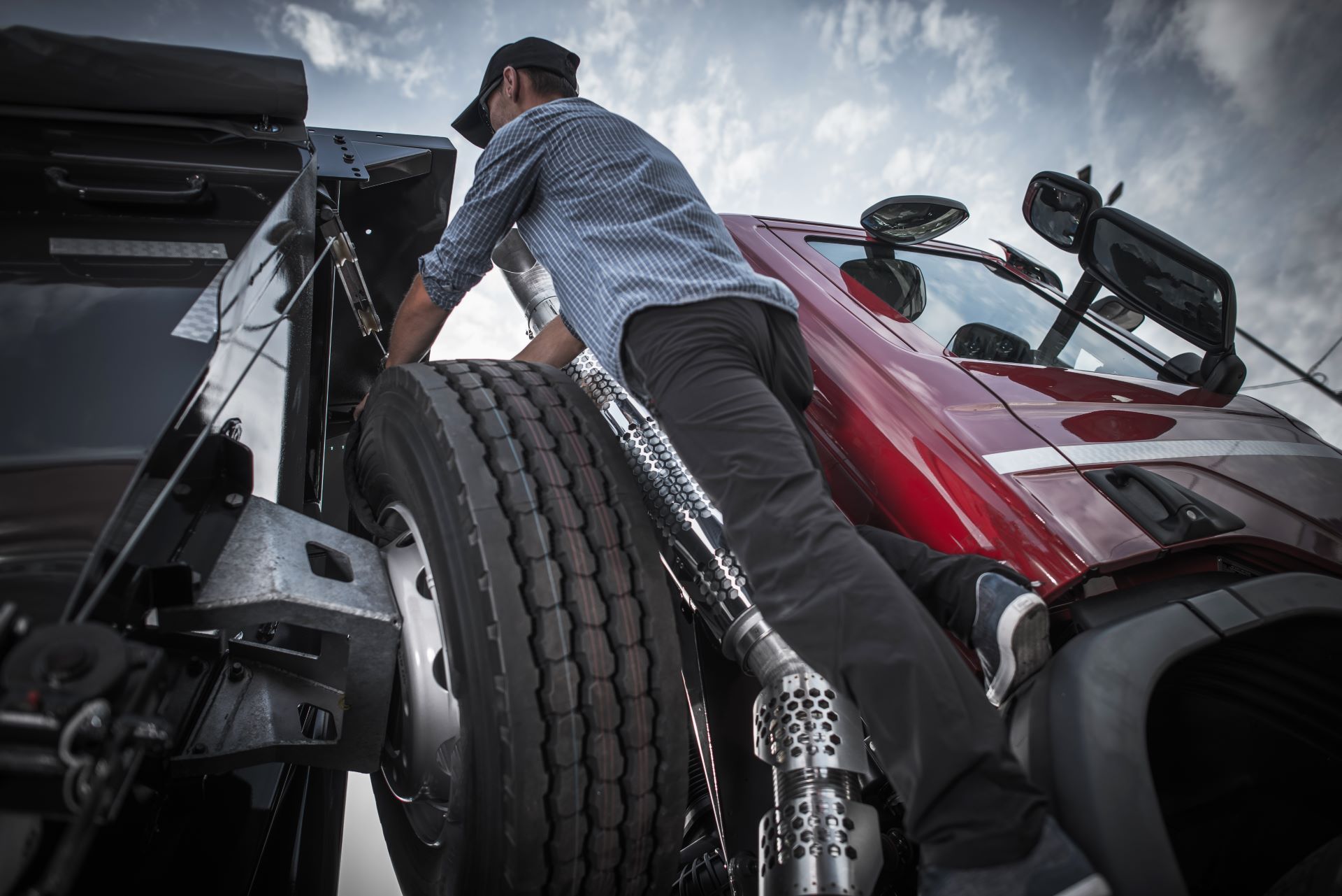Trucker inspecting semi-truck tire. Red cab, blue shirt, dark pants. Outdoors, cloudy sky.