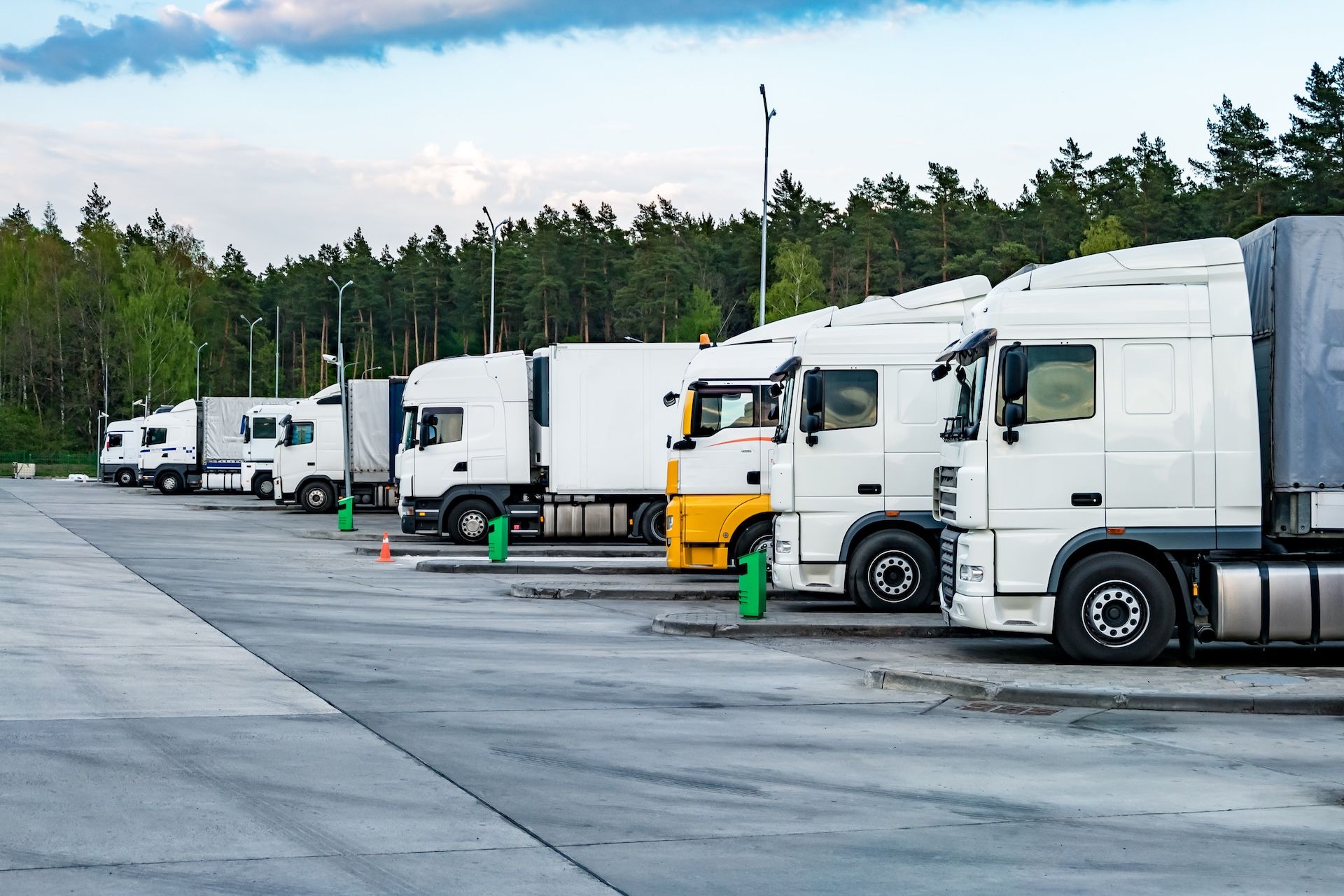 White semi-trucks parked in a line at a truck stop, set against a backdrop of trees under a cloudy sky.