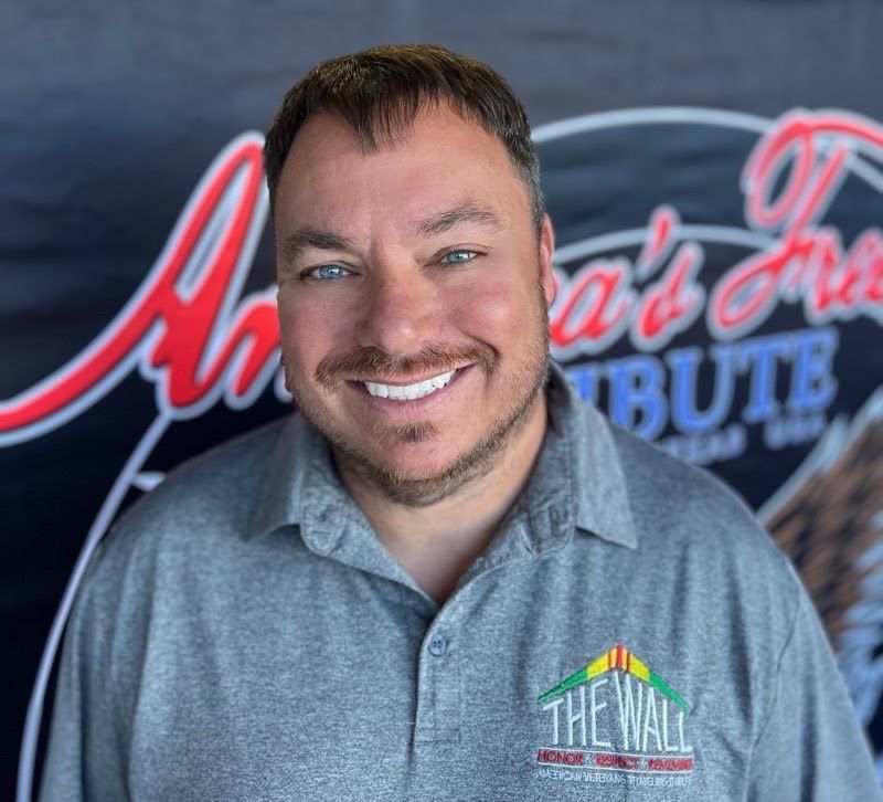 A man wearing a gray shirt that says the wall is smiling in front of a banner.