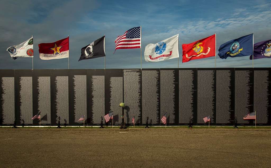 A row of flags are flying in front of a wall with names on it.