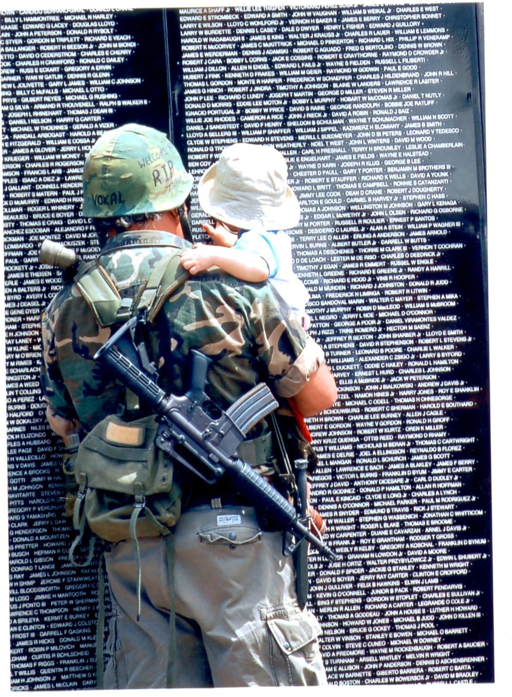 A soldier is holding a baby in front of a wall of names