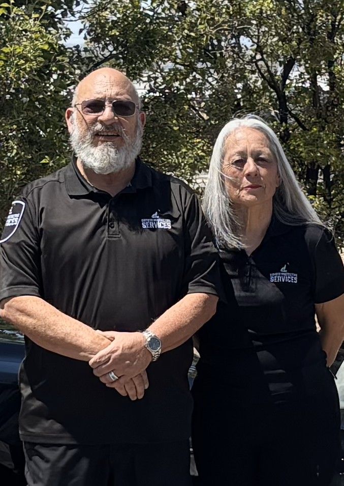 Man and woman in black polo shirts, standing side-by-side outdoors in front of trees, posing for the camera.