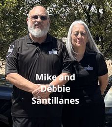 Mike and Debbie Santillanes in matching black shirts, smiling, standing in front of a car.
