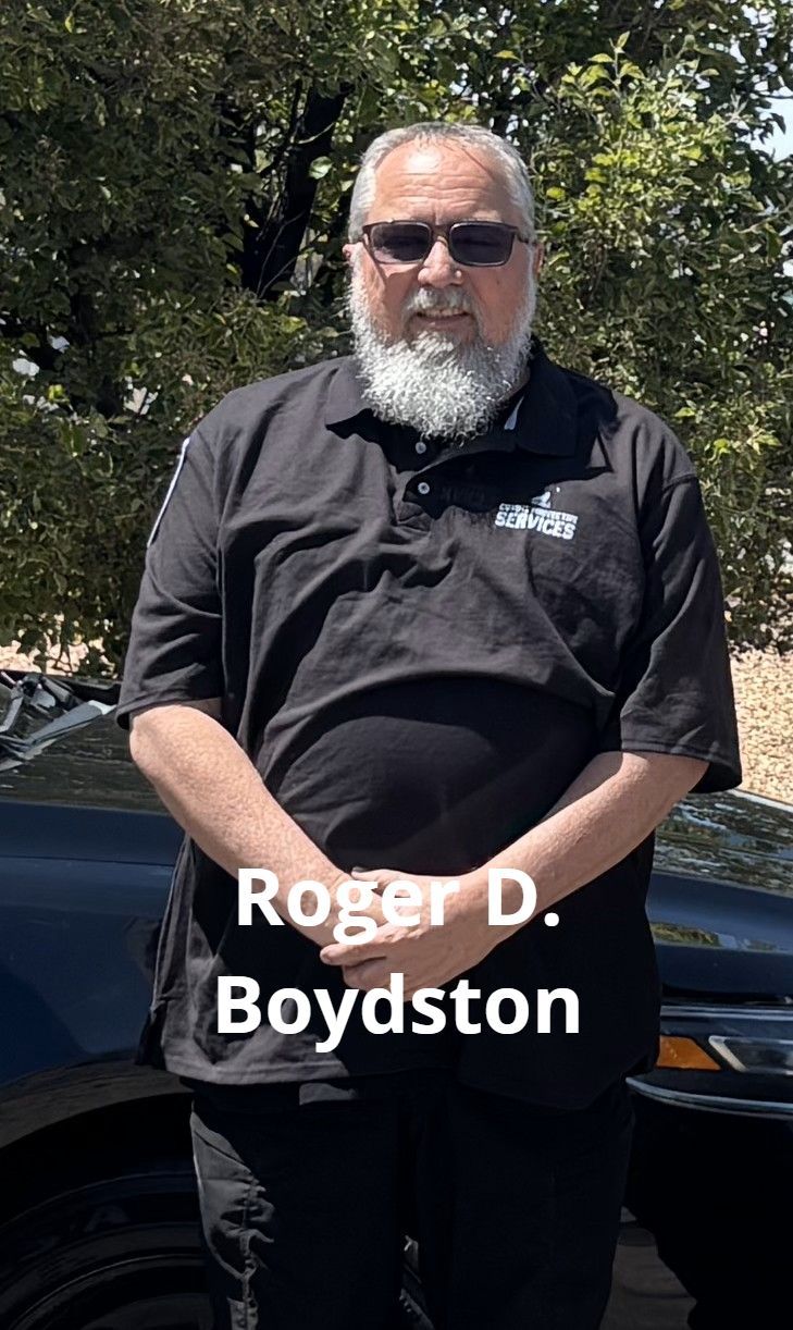 Roger D. Boydston stands outdoors, in a black polo shirt with a white beard, glasses, and a car behind him.