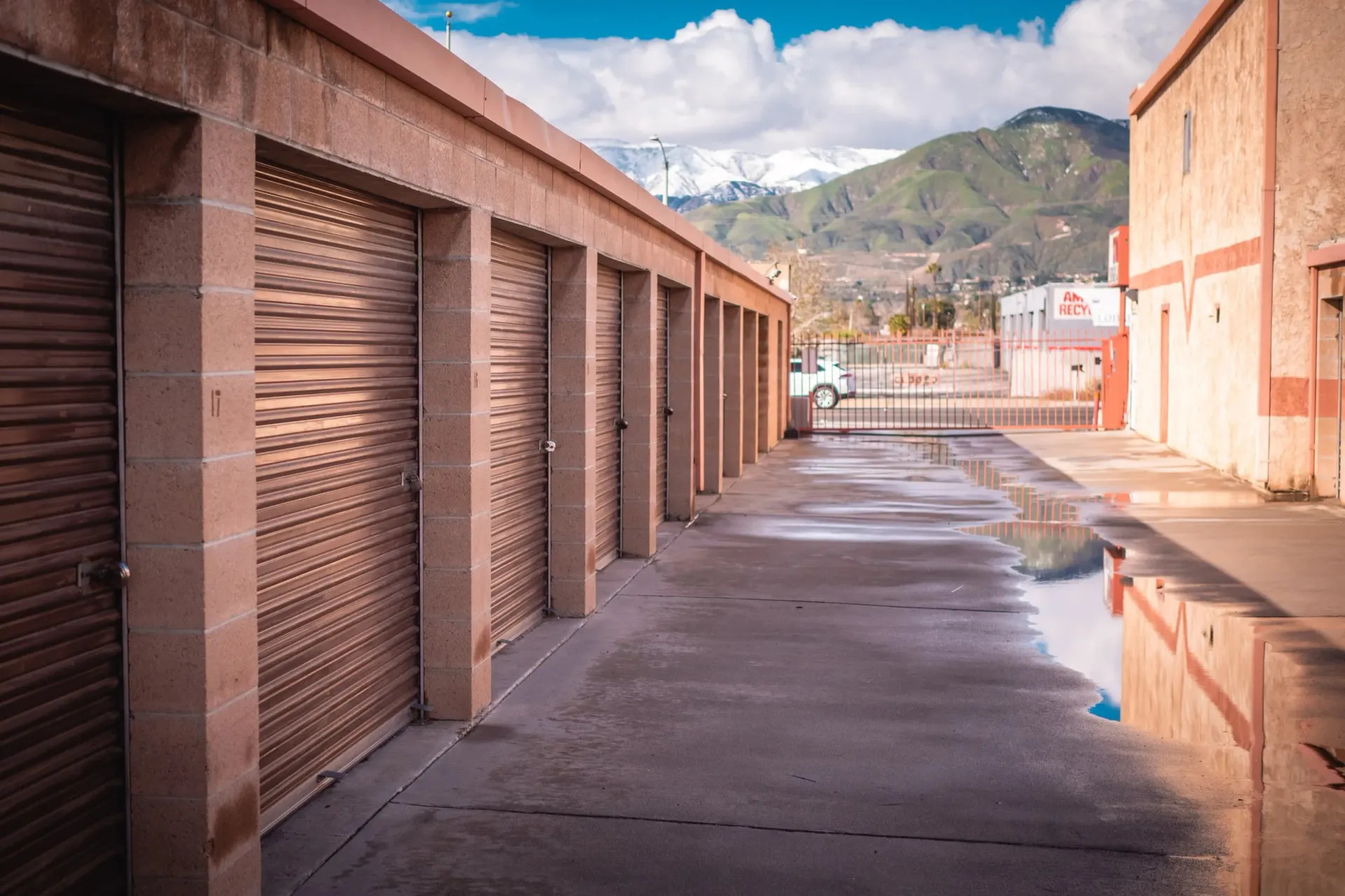 Storage units with roll-up doors line a concrete alley. Mountains and a partly cloudy sky are in the background.