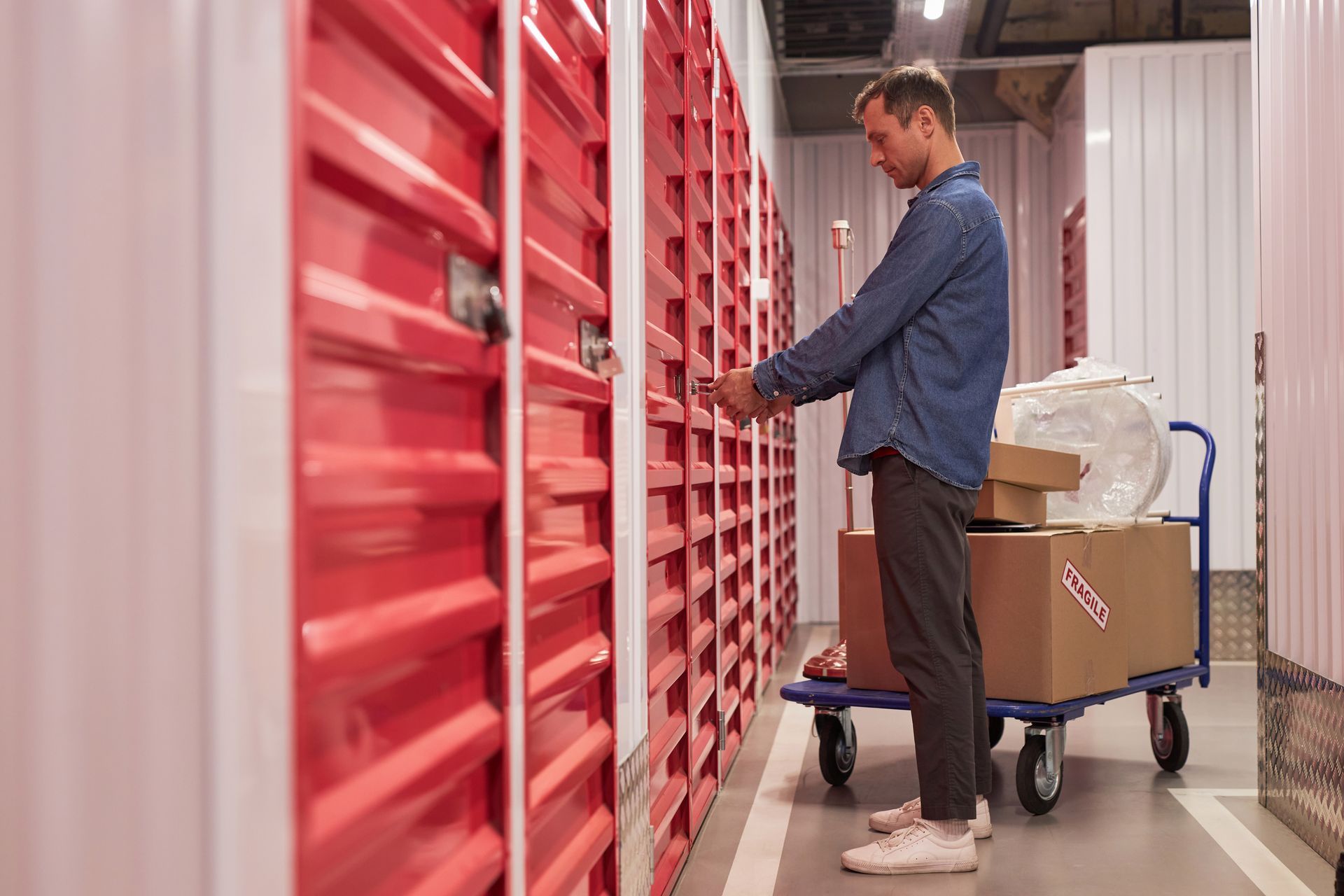 Man unlocking red storage unit door, boxes on a cart nearby. Interior storage facility.