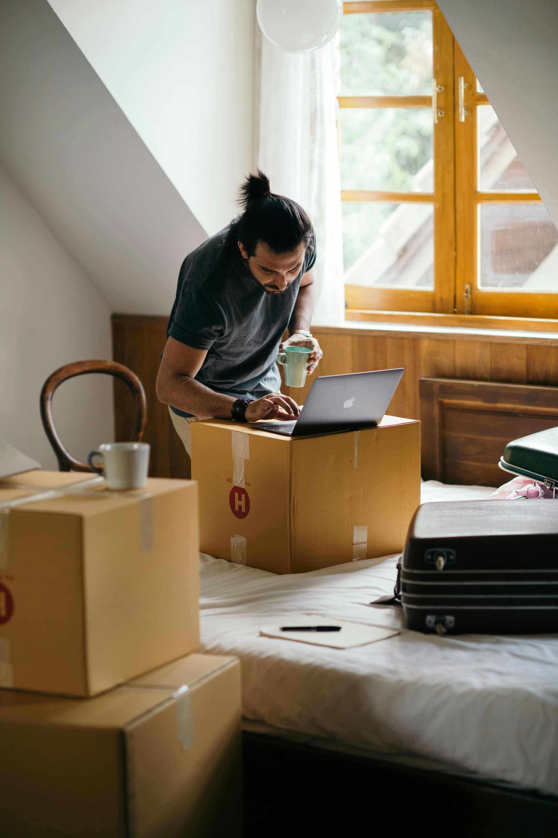Man using a laptop on a cardboard box in a bedroom, holding a mug. Boxes and a suitcase surround him.