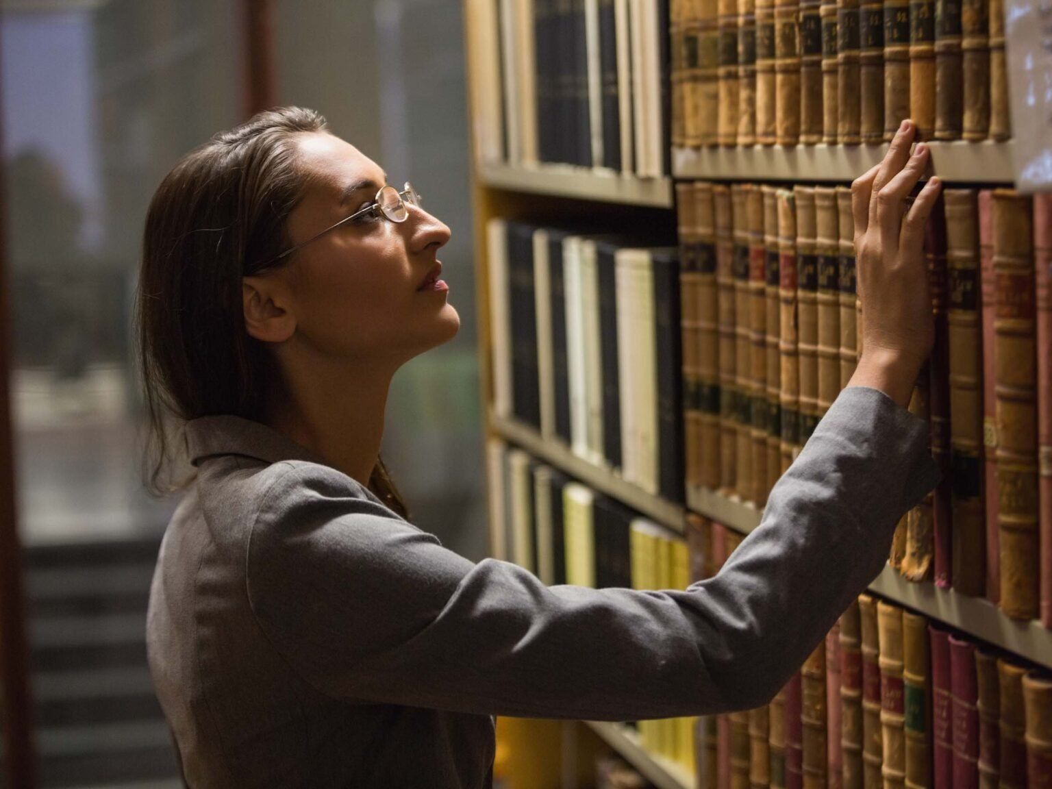 A woman is reaching for a book on a shelf in a library.