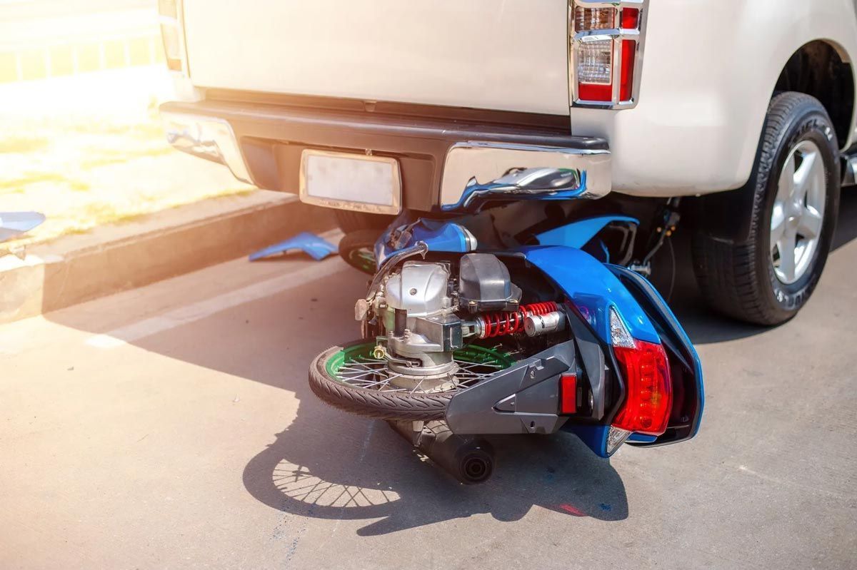 Blue motor scooter lies under white pickup truck after collision, showing visible damage on pavement