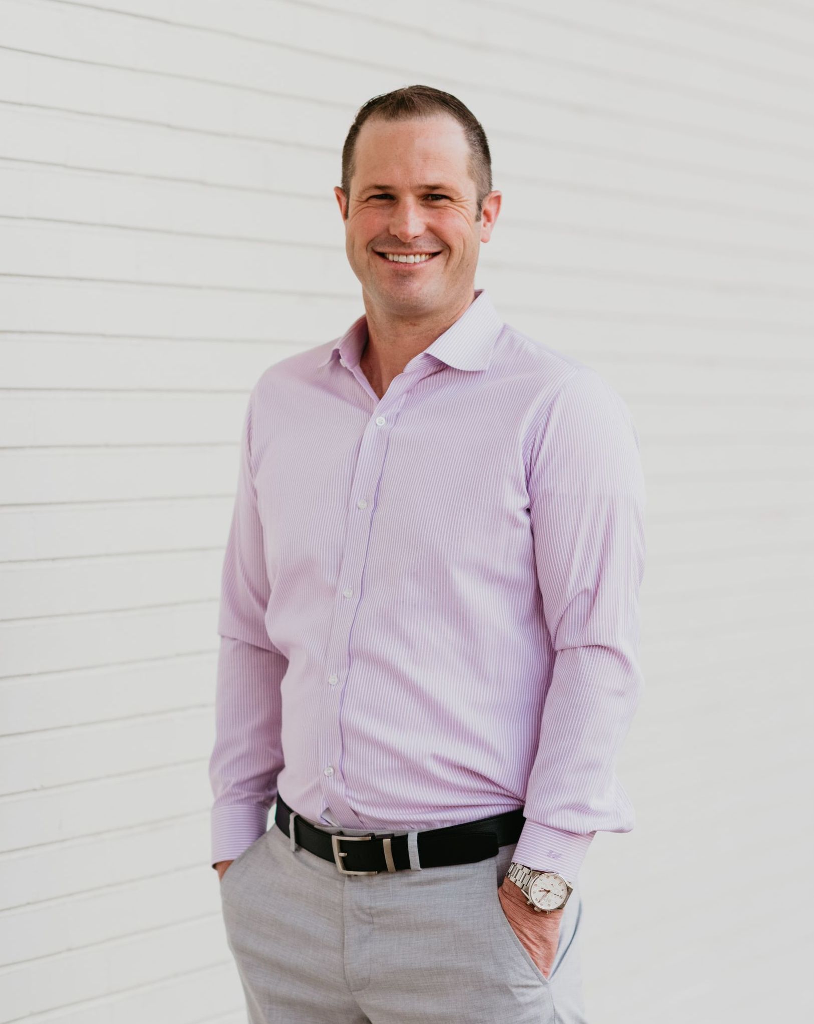 A man in a pink shirt and gray pants is standing in front of a white wall.