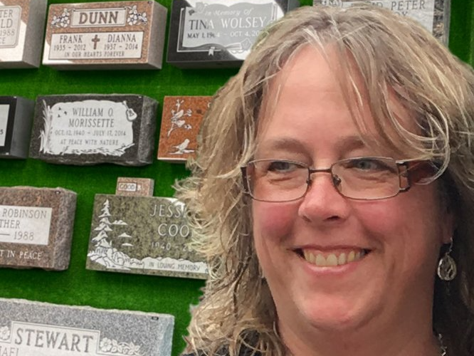 A woman wearing glasses is smiling in front of a wall of grave stones.