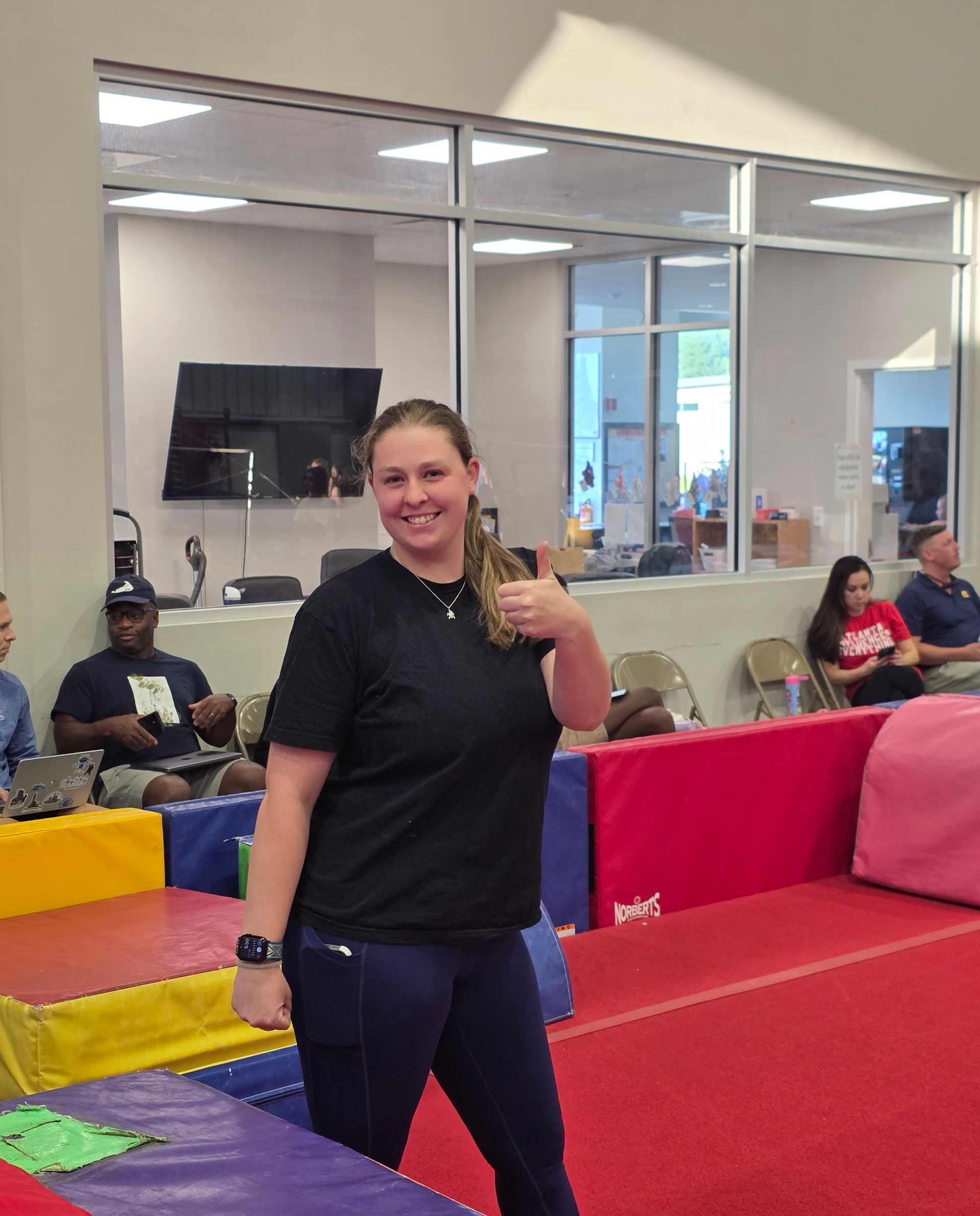 A smiling person in a black t-shirt and blue leggings gives a thumbs up inside an indoor gymnastics training facility.