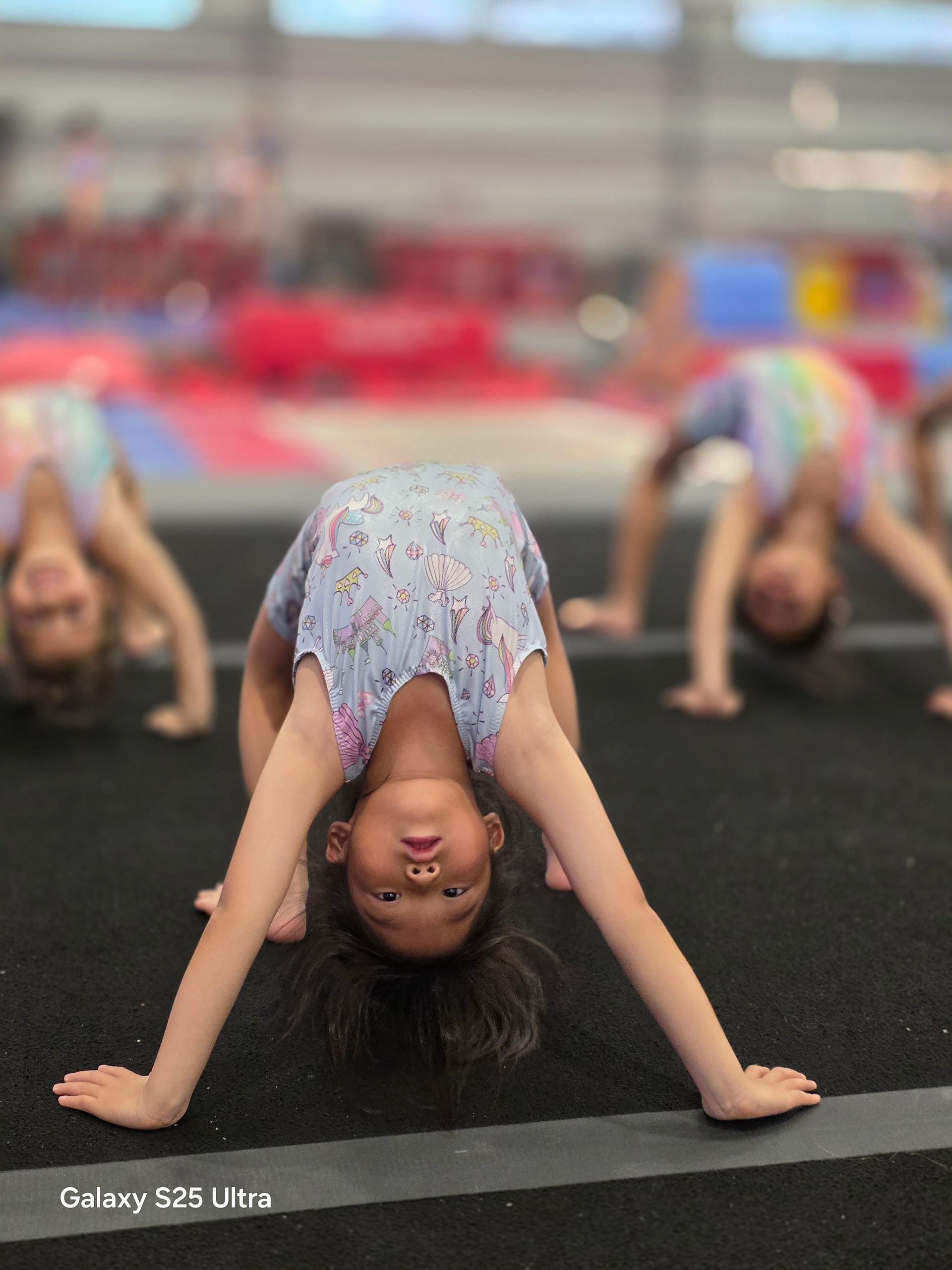 Three children perform backbends on a black gym mat in a training facility.