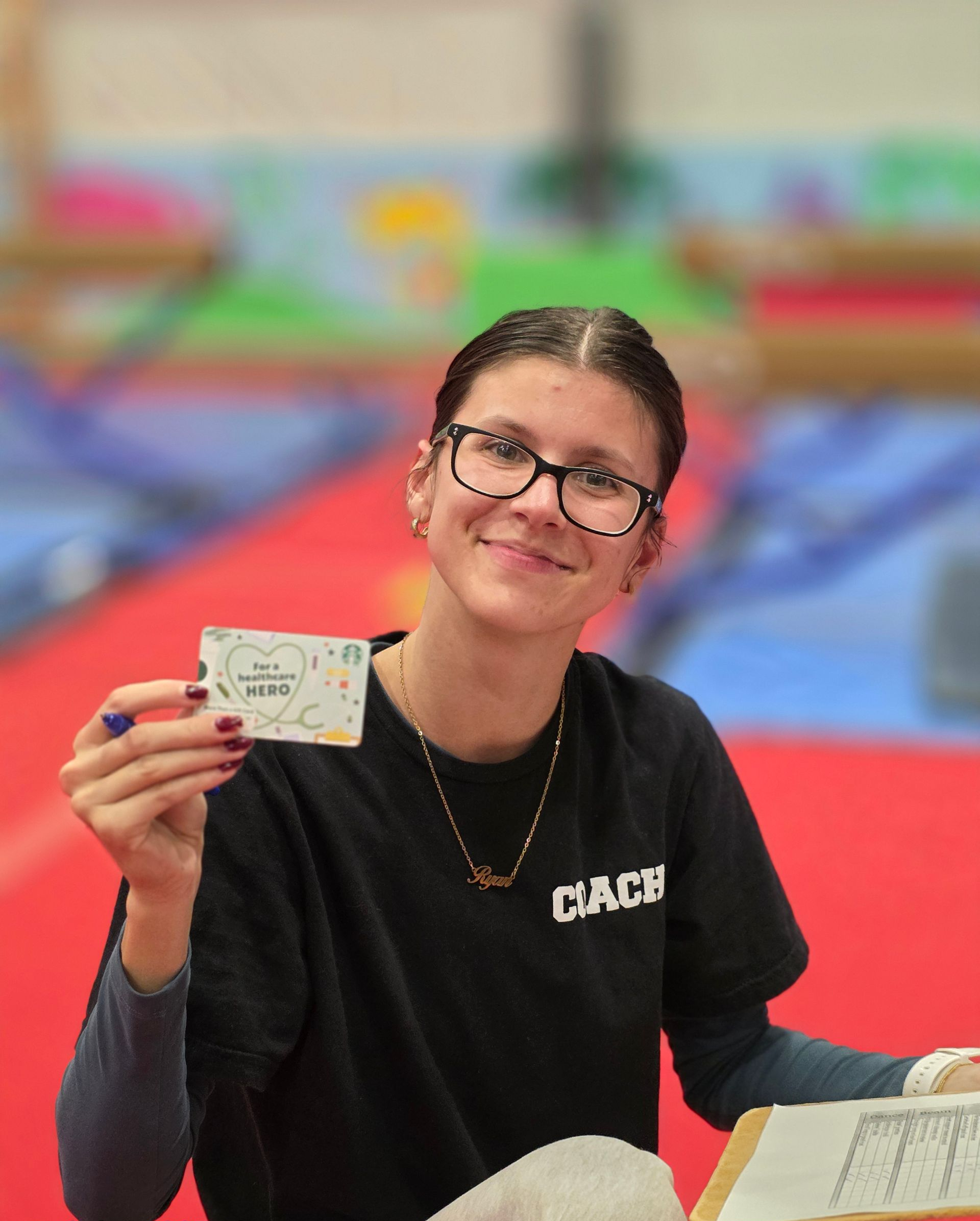 A coach smiling while holding a small gift card in a gym, with sports equipment visible in the background.