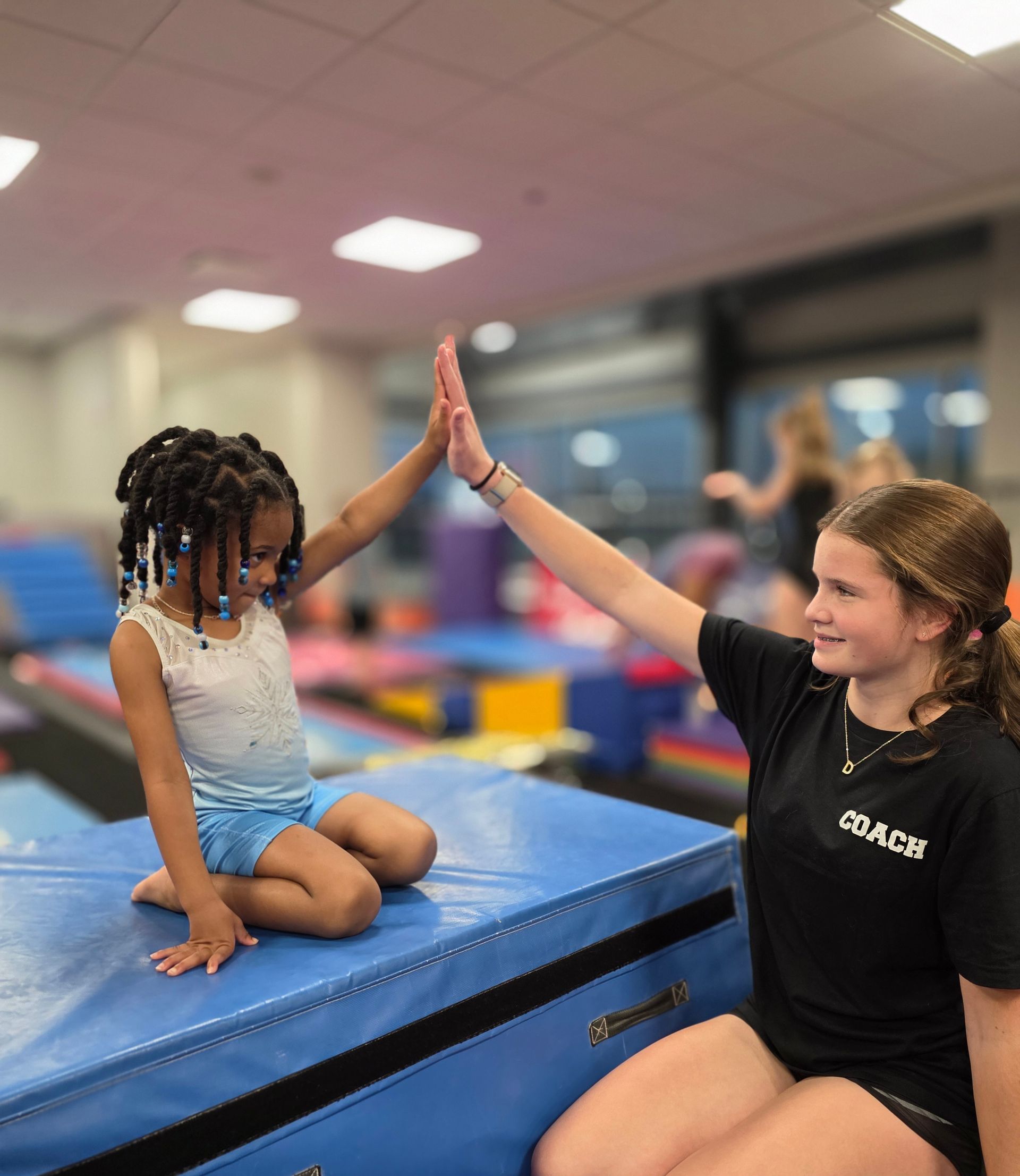 A young gymnast and a coach in a black t-shirt exchange a high-five while sitting on a blue mat in a gym.