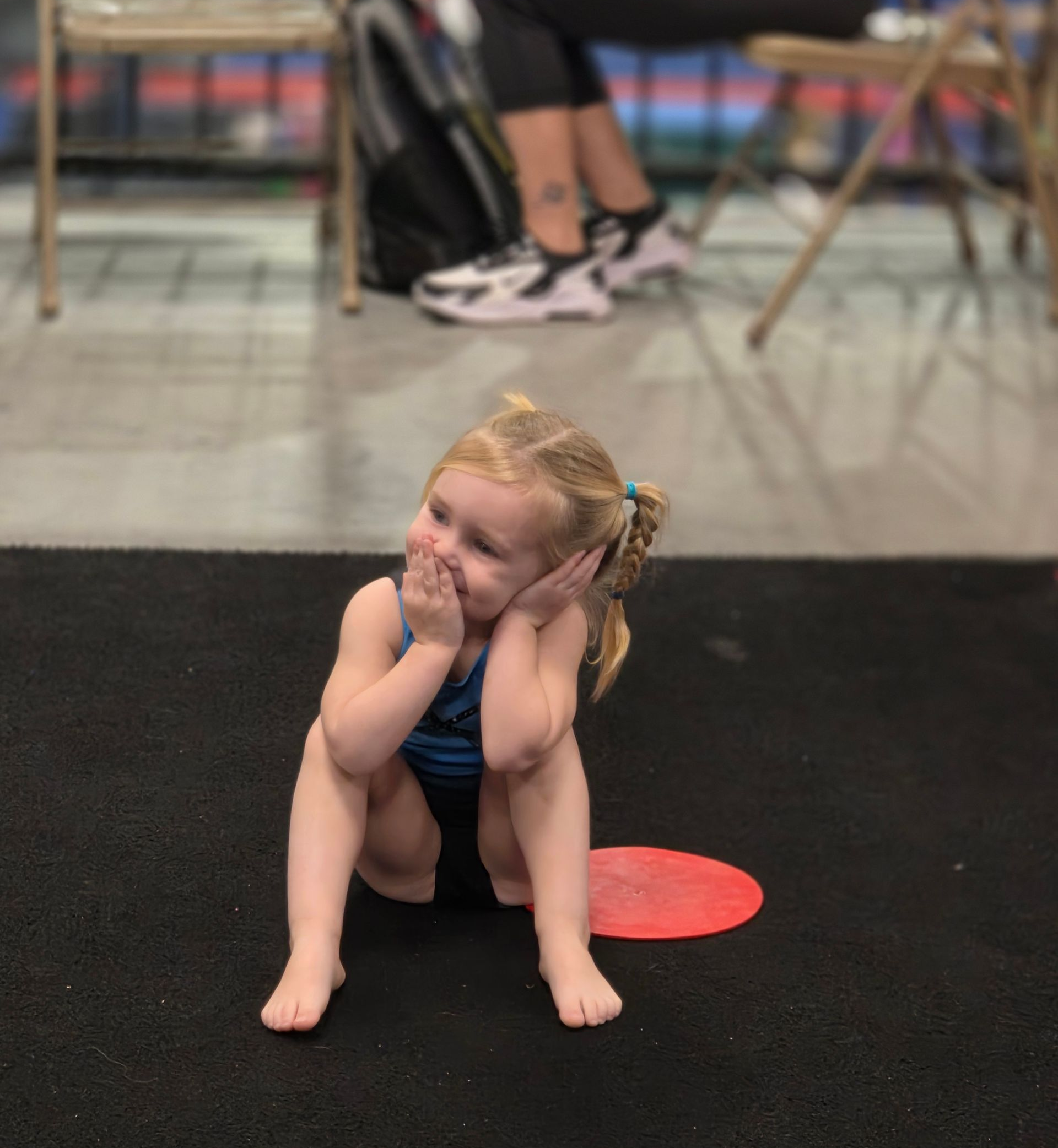 A child in a blue leotard sits on a black mat with their hands on their face, looking to the side.