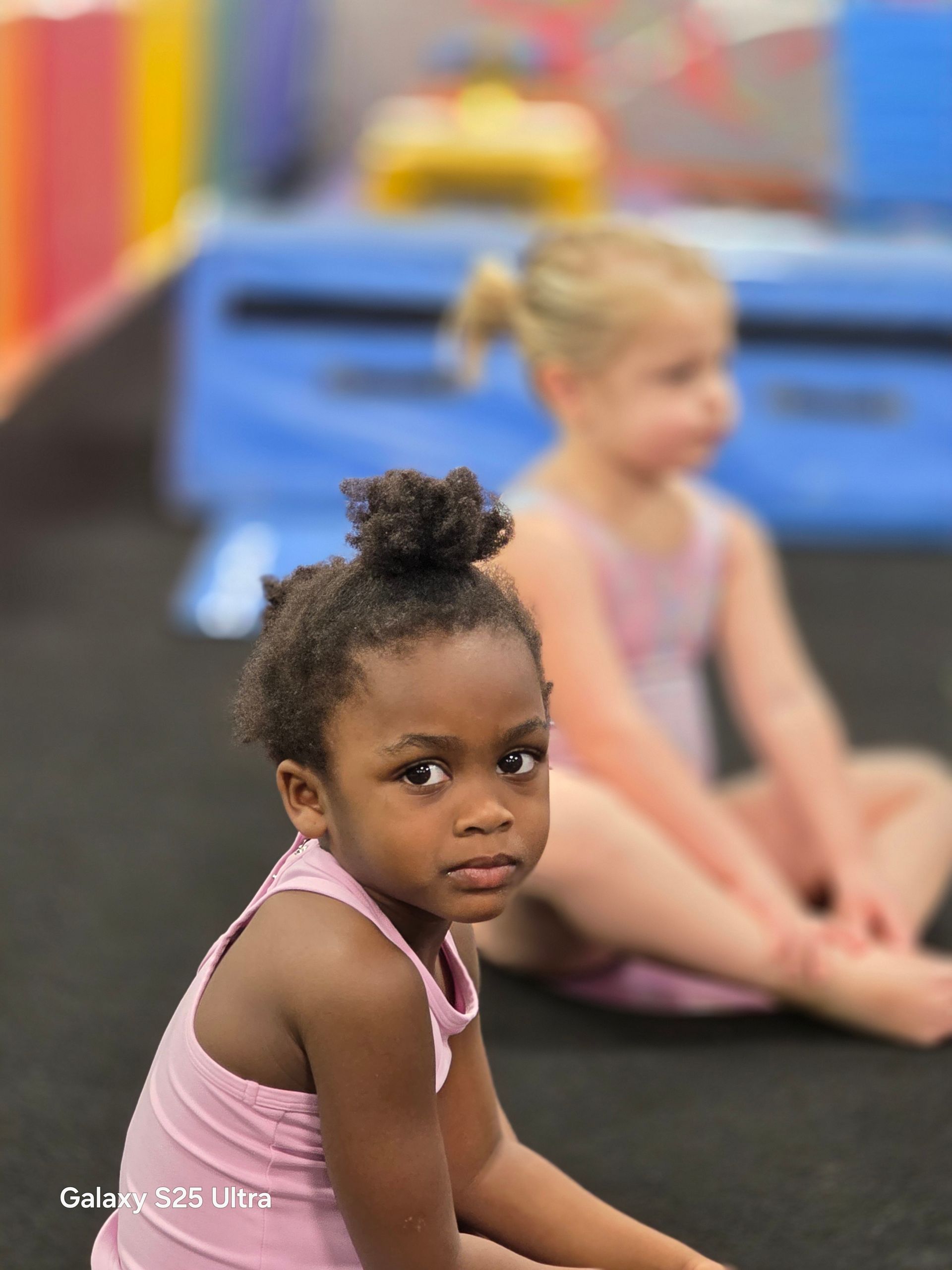 Two children in pink leotards sit on a black floor in an indoor gym with colorful equipment in the blurred background.