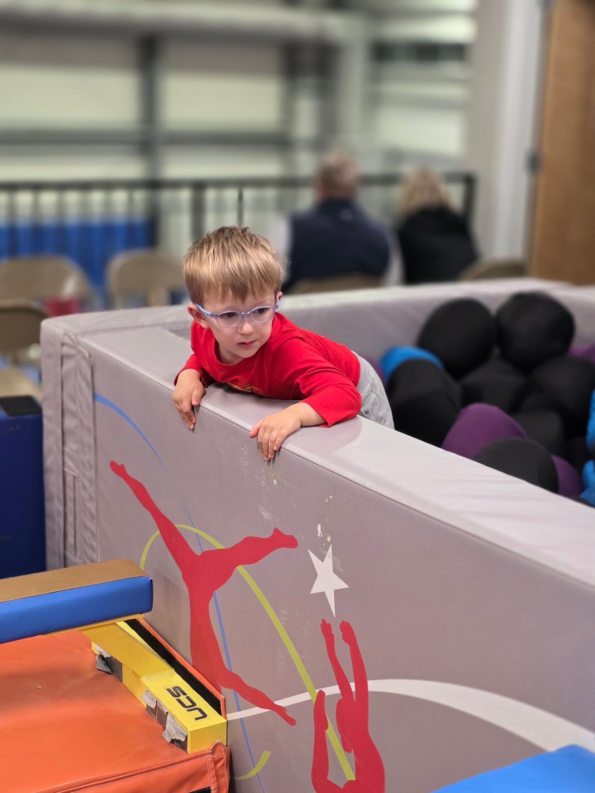 A young person in a red long-sleeved shirt leans over a gym mat decorated with red silhouettes, looking toward a foam pit.