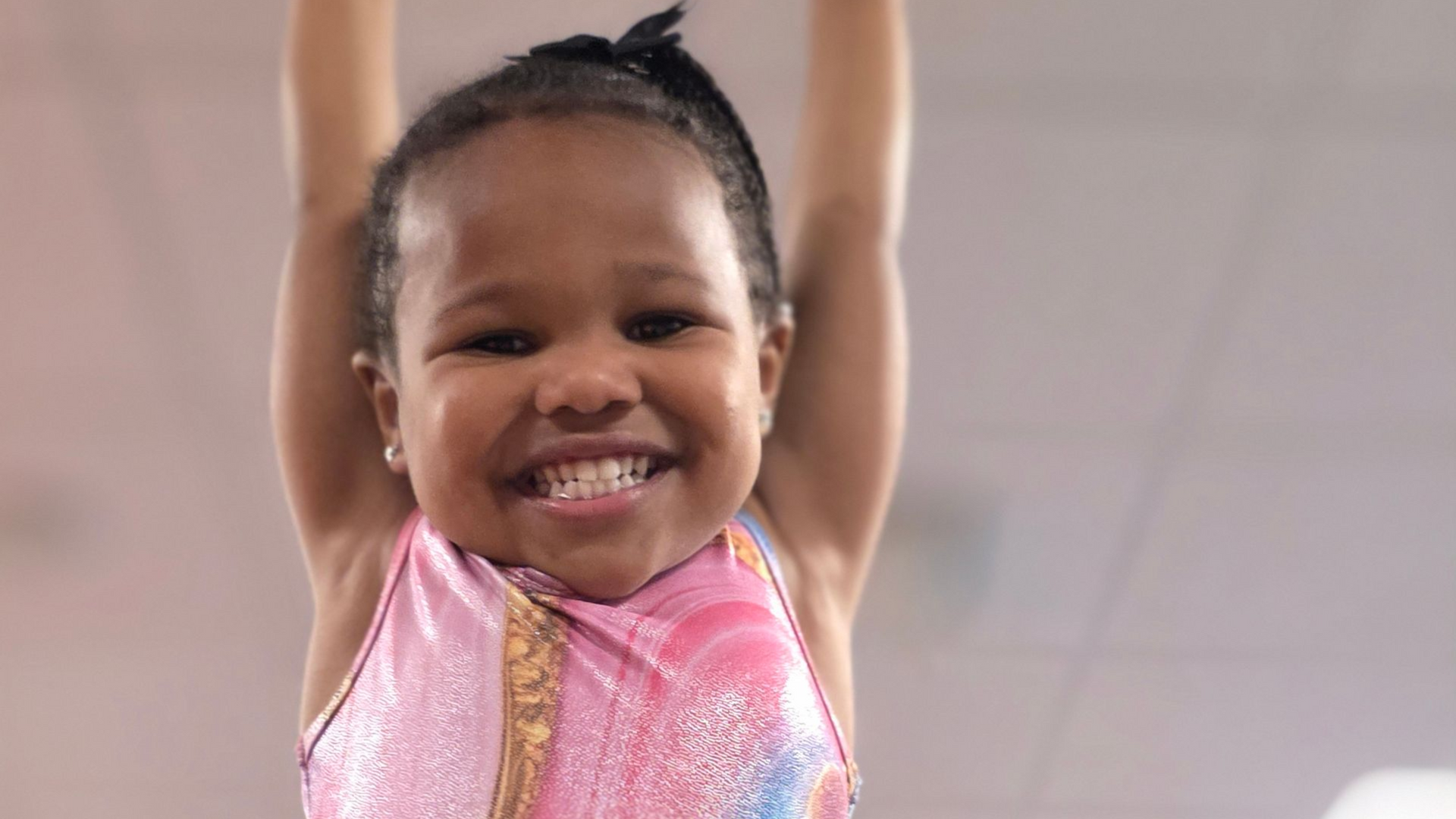 A smiling child in a pink gymnastics leotard hangs with their arms raised high against a plain, light background.