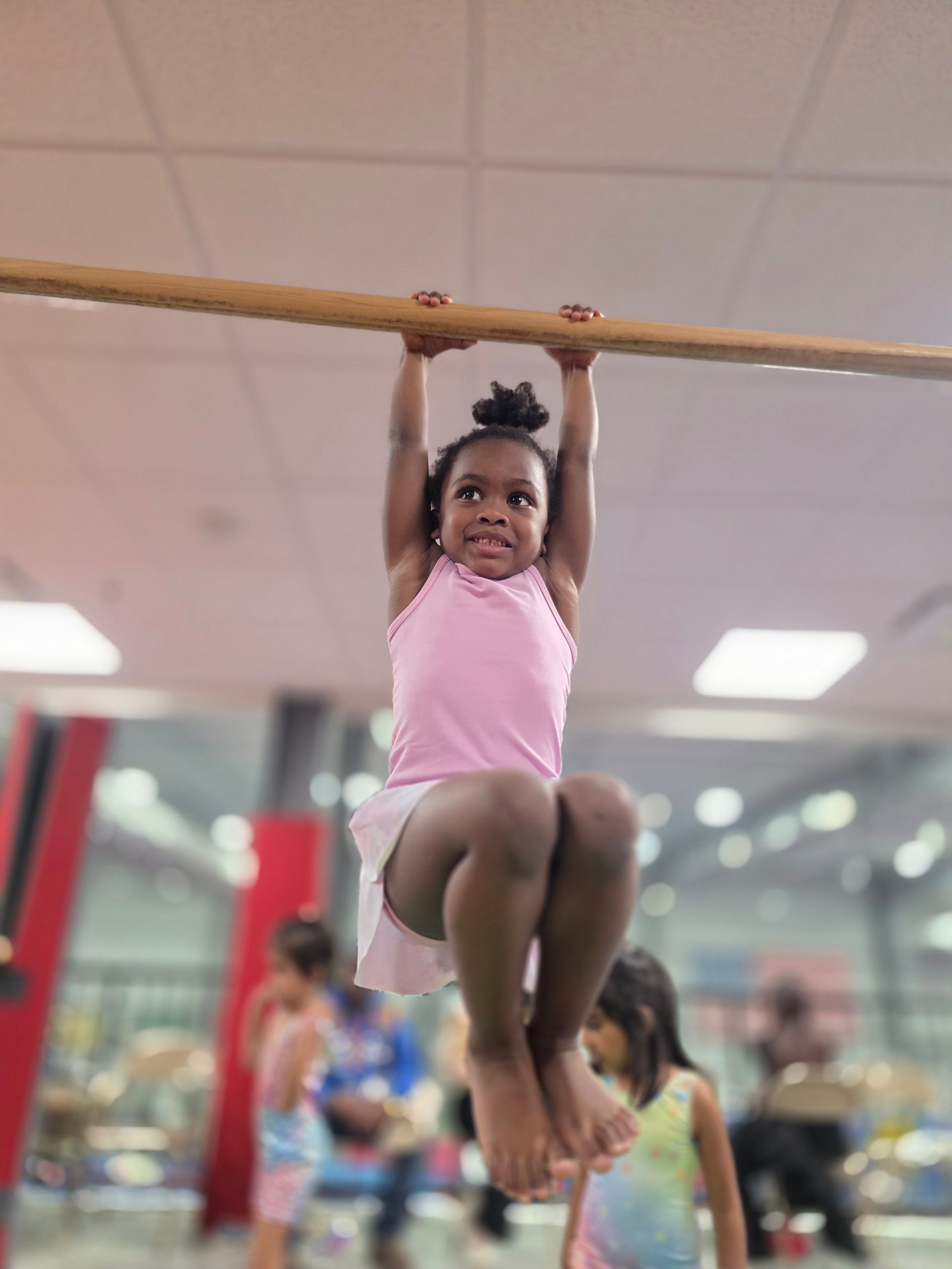 A child in a pink shirt and skirt hangs from a wooden bar in a gymnastics studio.