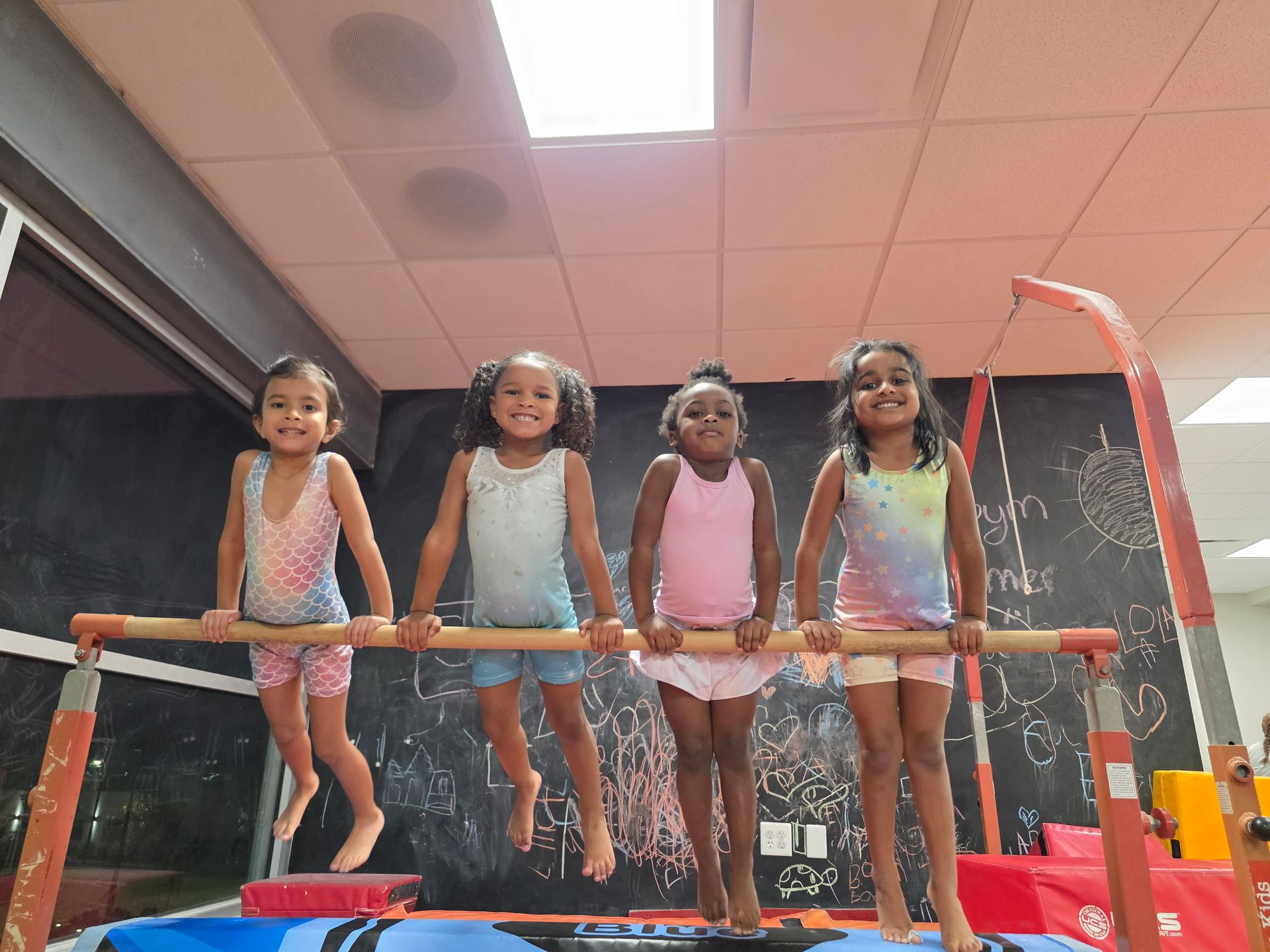 Four children in athletic wear smile while hanging from a low gymnastics bar in a play area.