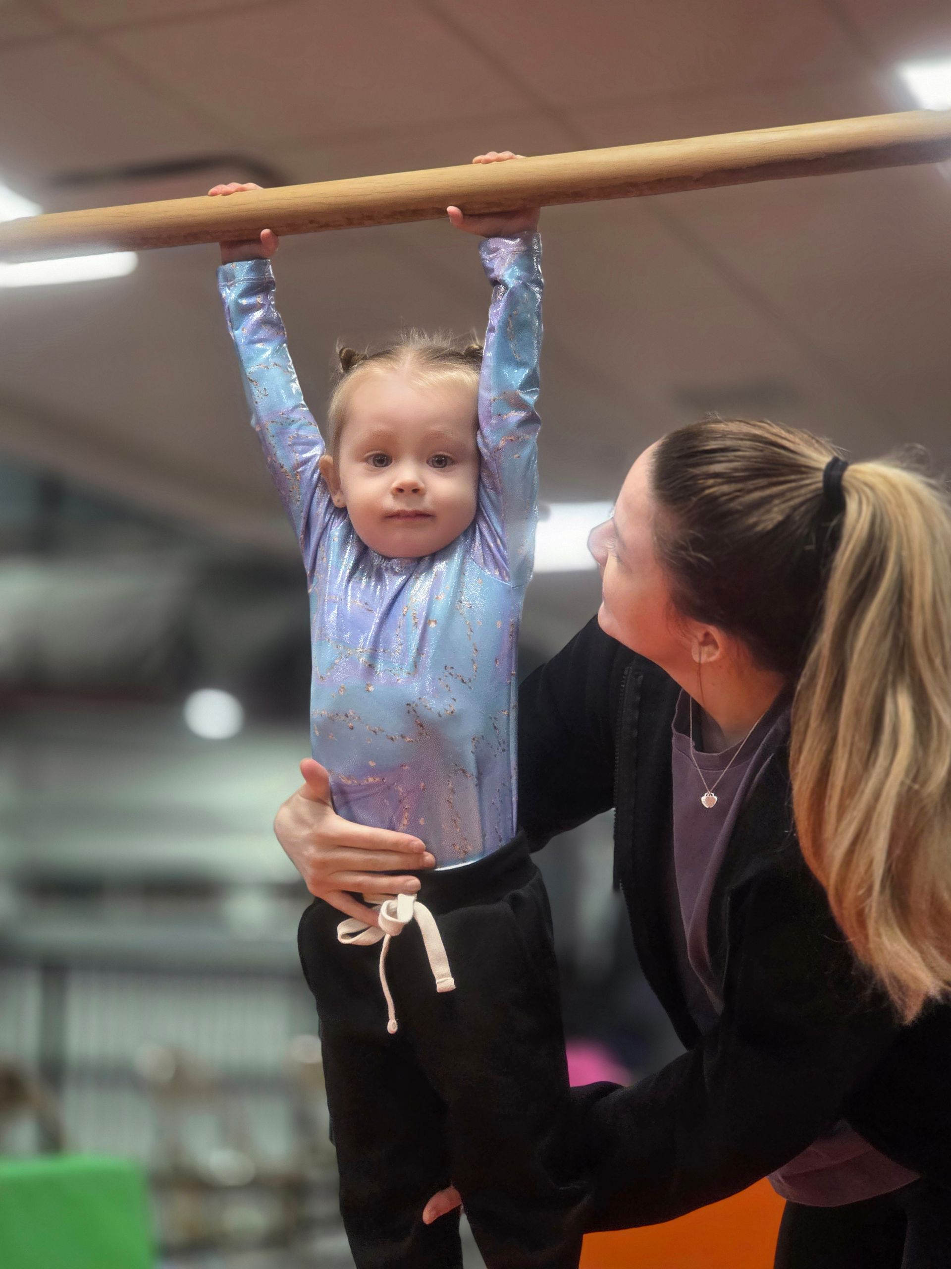 A coach supports a child wearing a sparkly blue leotard as they hang from a wooden gymnastics bar in a gym.