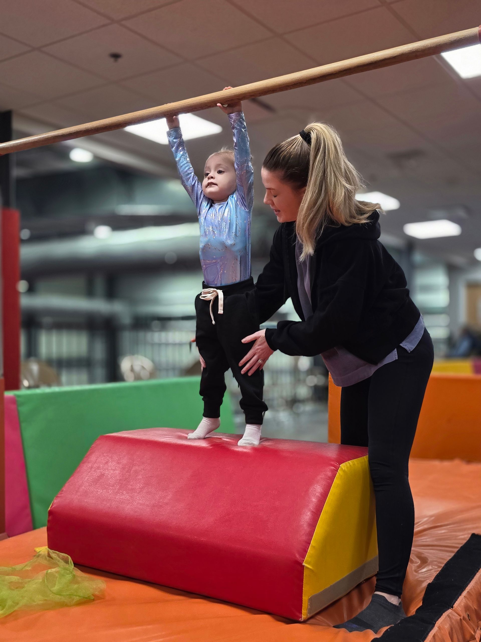 A person helps a young child hang from a horizontal bar while standing on a red and yellow gymnastics foam block.