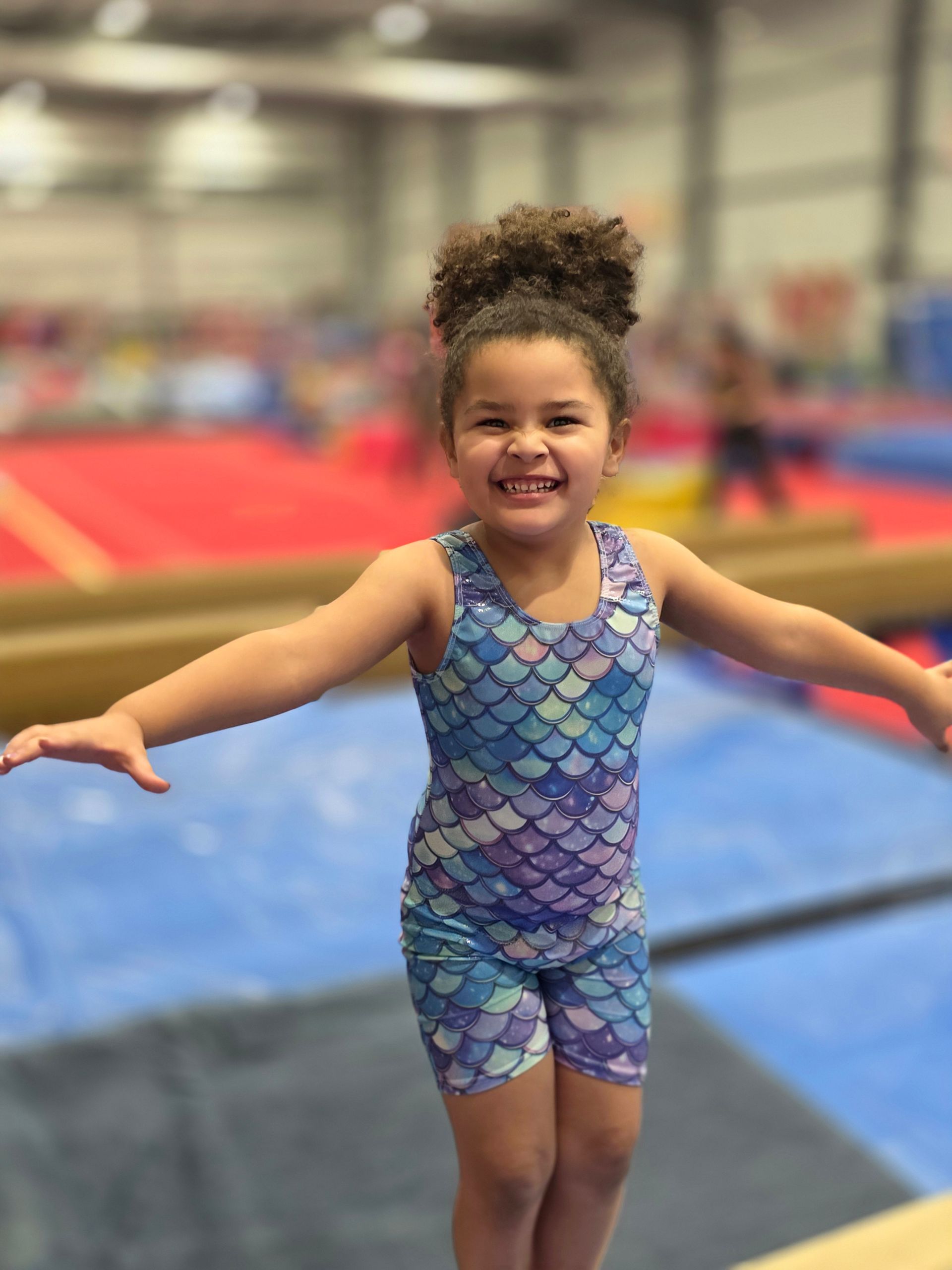 A child wearing a blue and purple scale-pattern leotard smiles with arms outstretched while practicing on a balance beam.