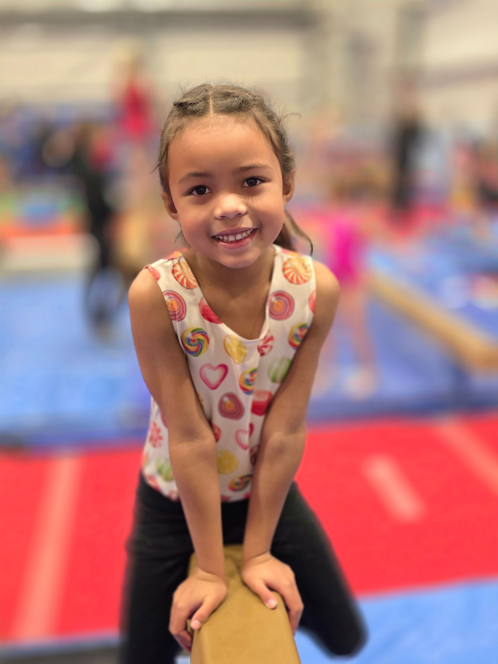A smiling child in a colorful patterned leotard balances on a gymnastics beam inside a gym.