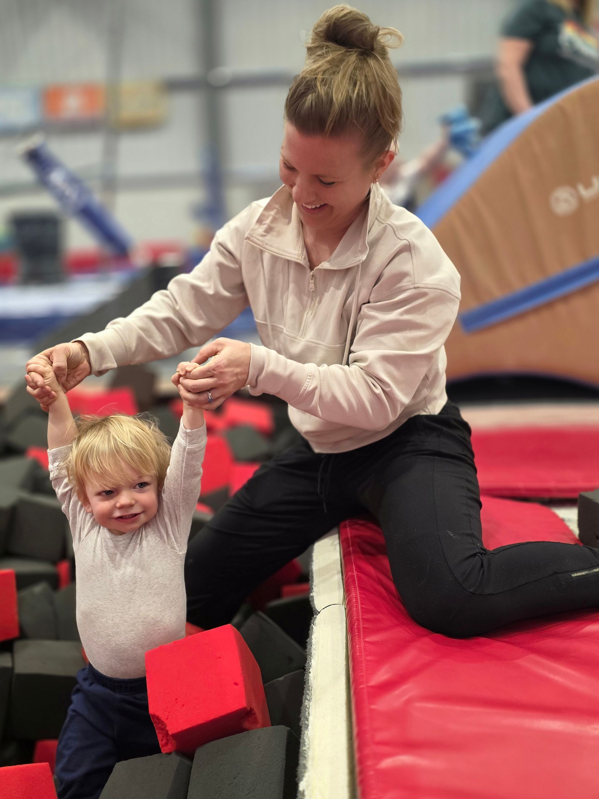 An adult holds a child’s hands to help them stand in a foam pit at an indoor gymnastics center.