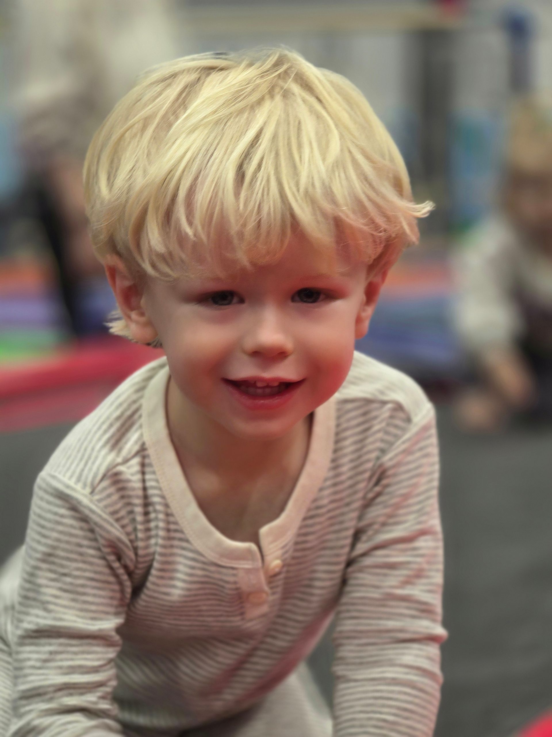 A smiling child with blonde hair wearing a long-sleeved, light-colored striped shirt, indoors on a mat.