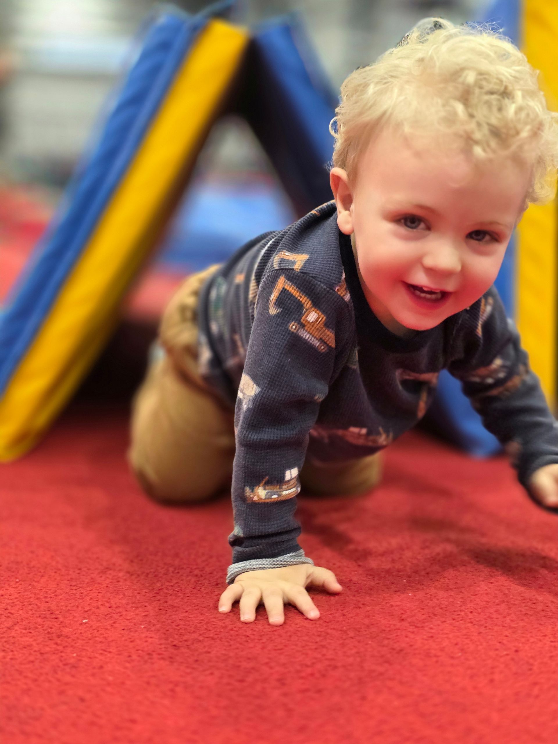 A curly-haired toddler smiling while crawling through a triangular blue and yellow foam tunnel on a red carpeted floor.