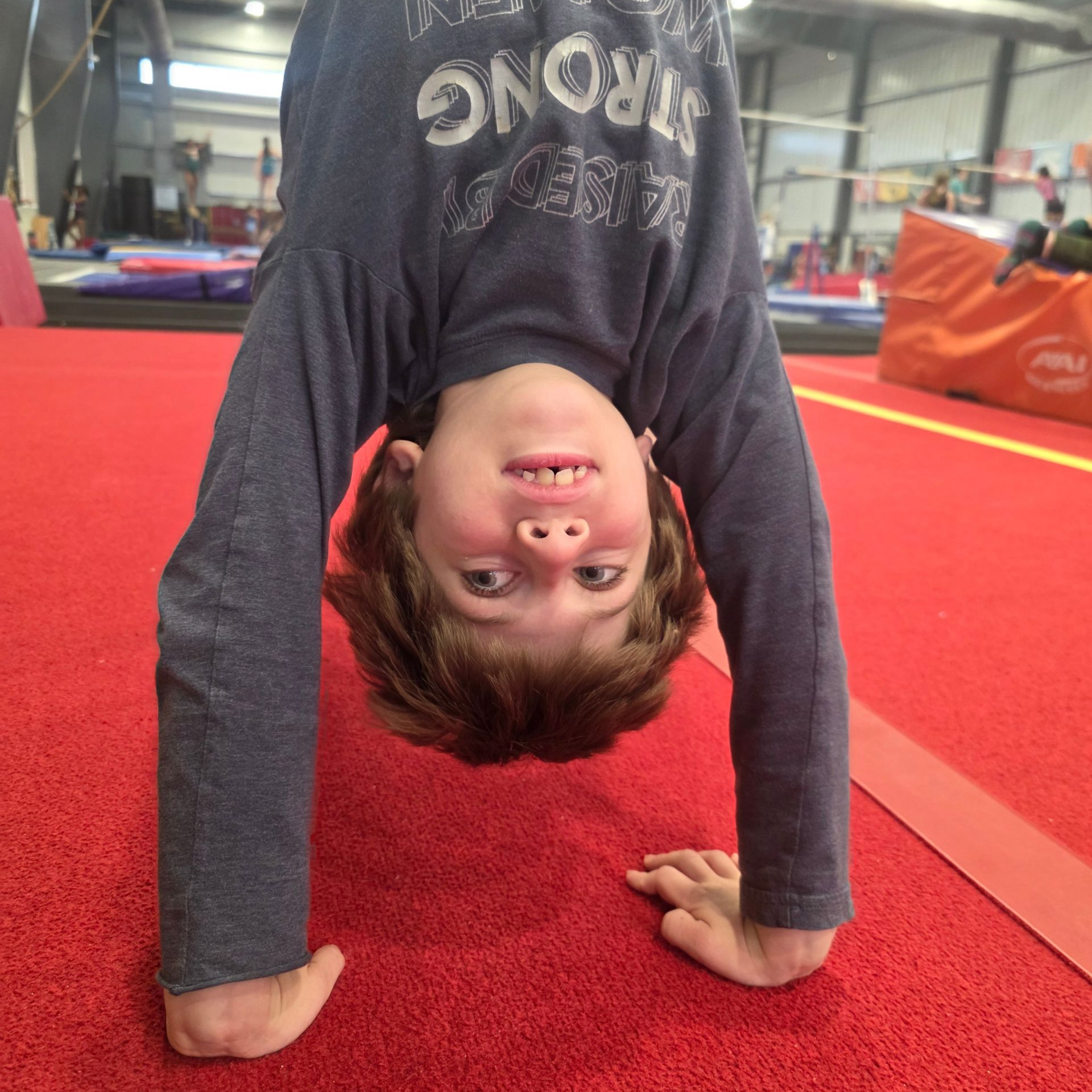 A person in a gray long-sleeved shirt doing a handstand on a red gym floor.