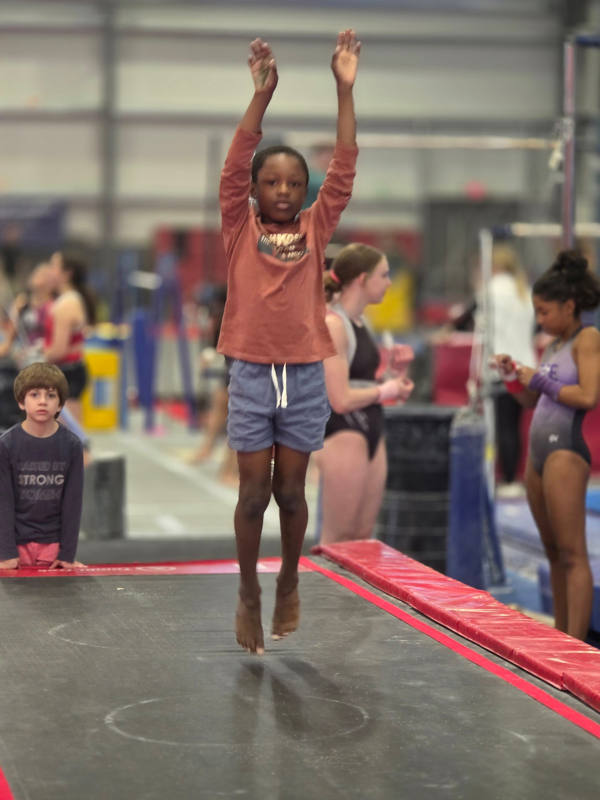 A person in a rust-colored long-sleeve shirt and blue shorts jumps high on a trampoline inside a gymnastics gym.