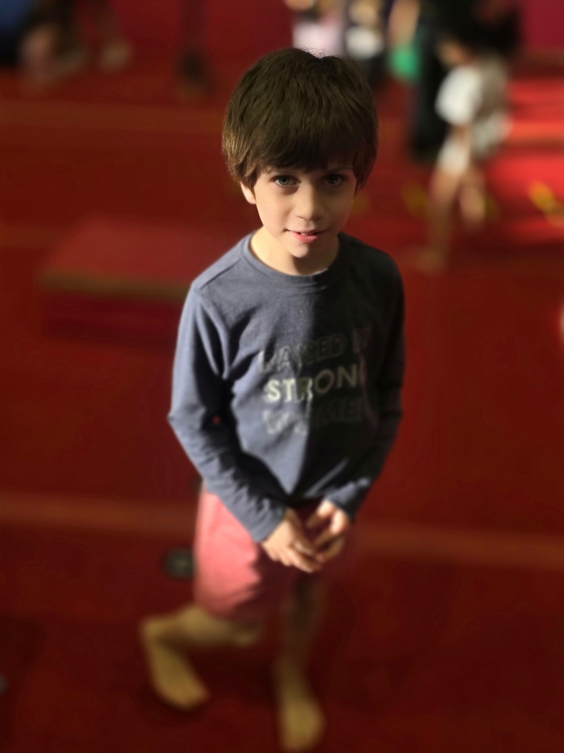 A young child in a long-sleeved gray shirt and red shorts stands on a red mat in a gymnasium.