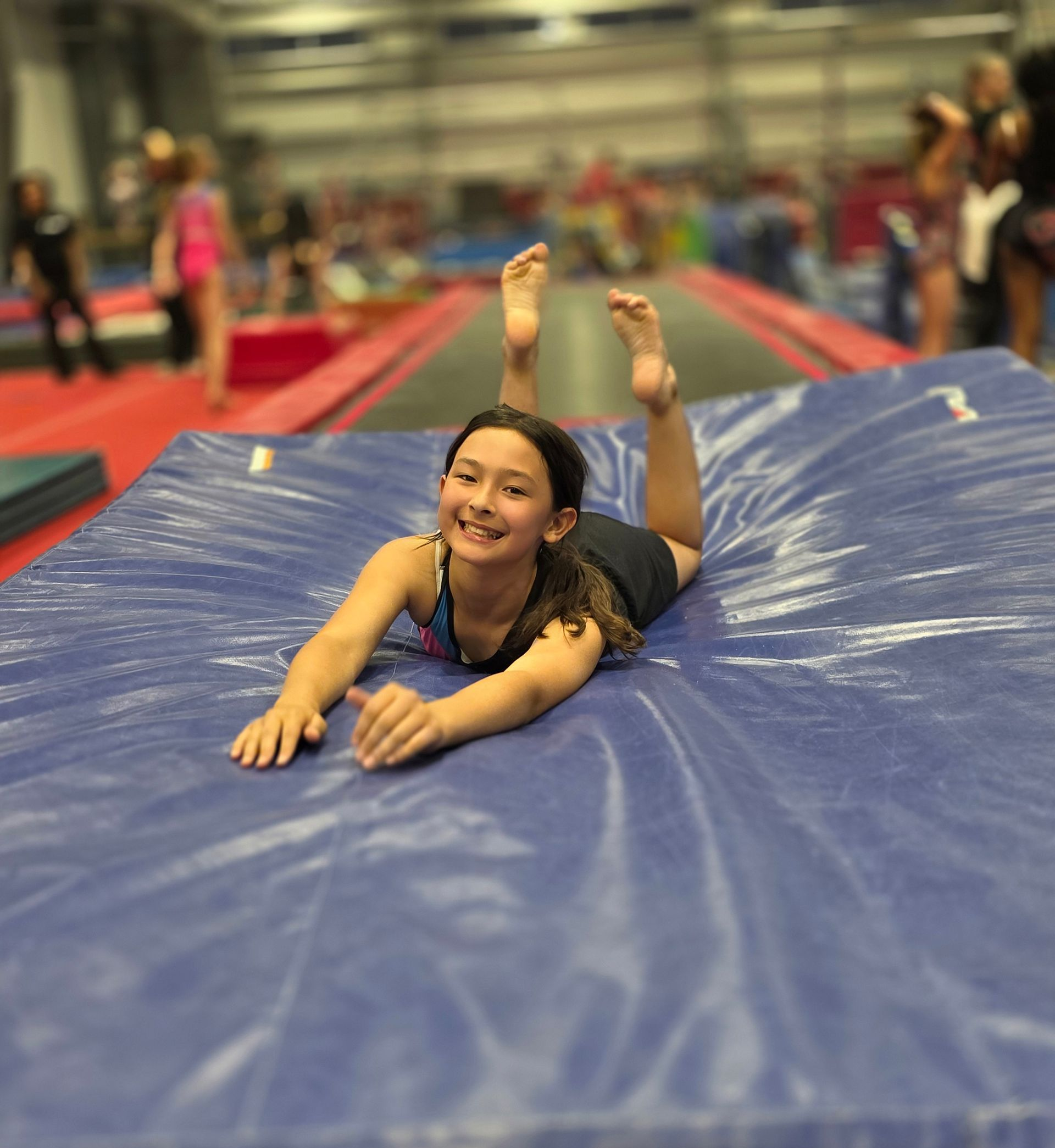 A person lying on their stomach on a blue gym mat, smiling, with feet raised in the air inside a training facility.