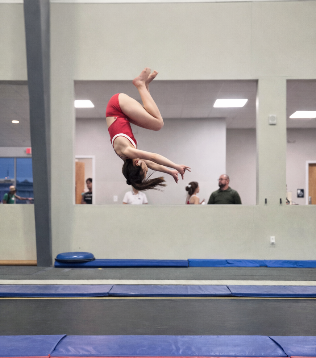A gymnast in a red leotard performs a back tuck on a trampoline in a gym.