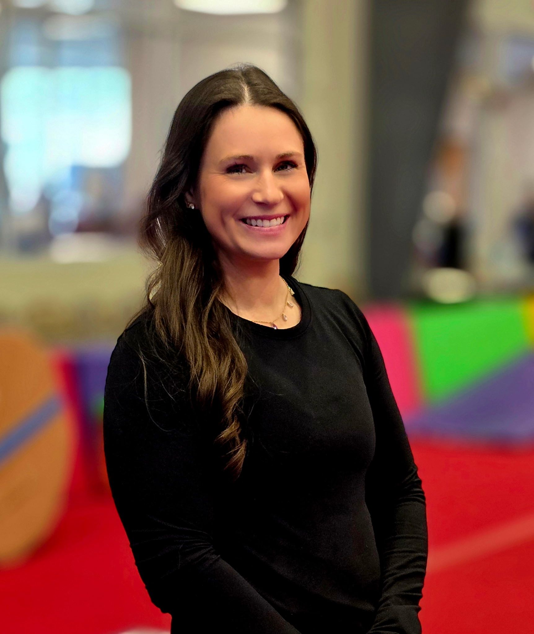 A smiling person with long dark hair, wearing a black long-sleeved shirt, stands in front of a colorful, blurred background.