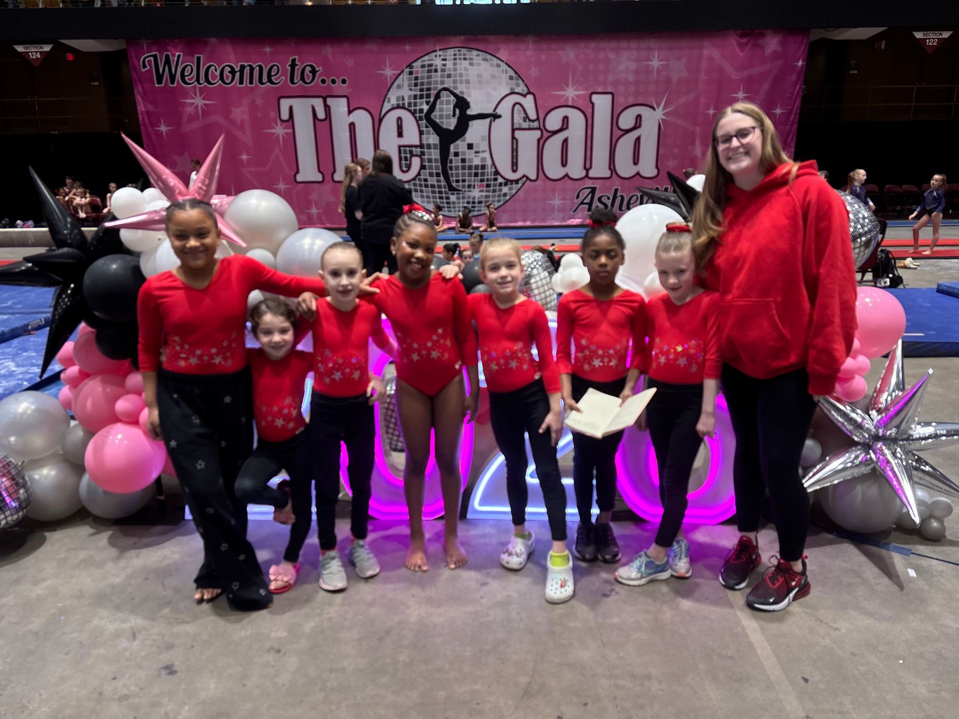 A group of children and a leader in red shirts stand before a balloon display and a sign for The Gala at a gym.